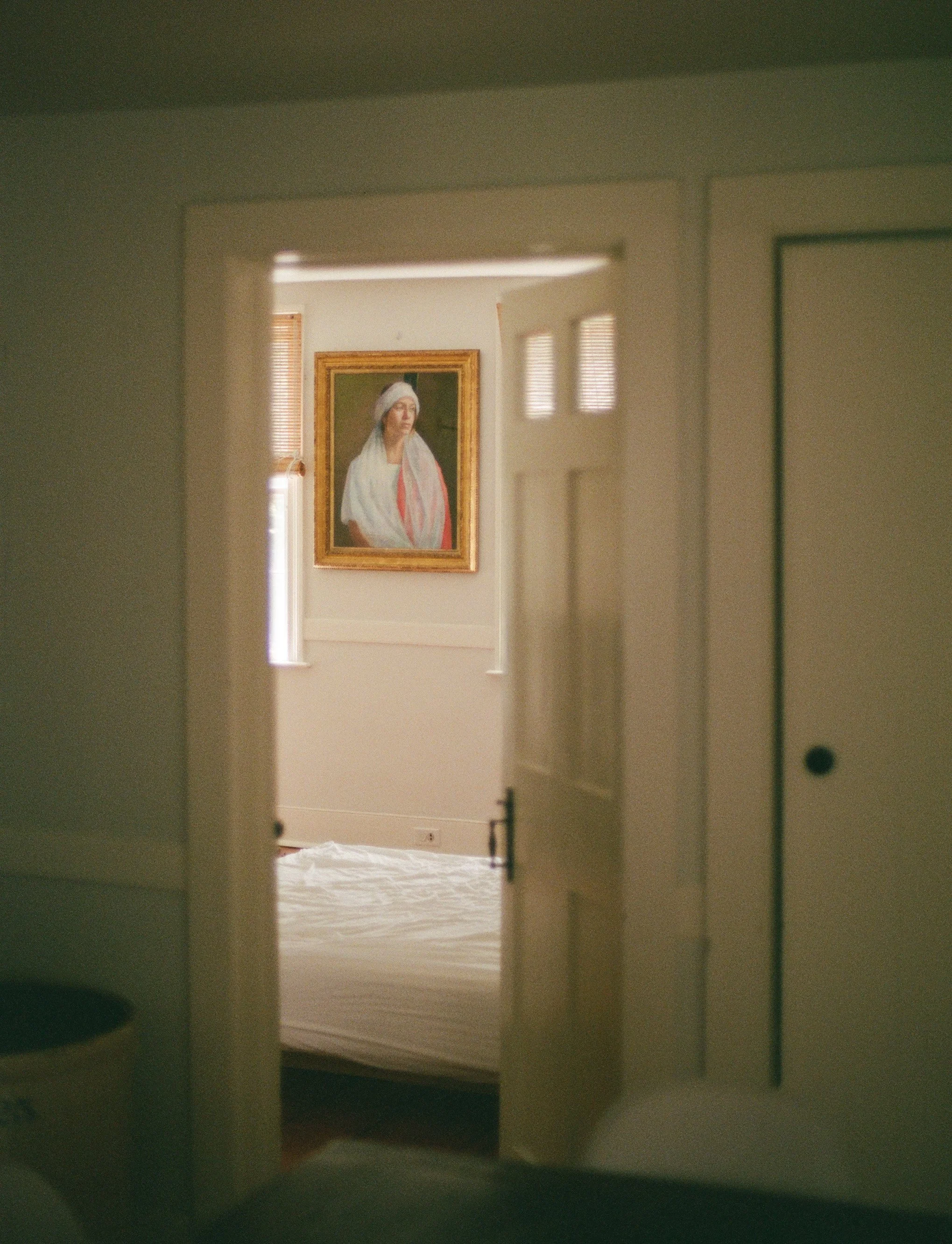 Photo of a bedroom viewed through a doorway, featuring a portrait of a woman in a white headscarf and pink sash on the wall.