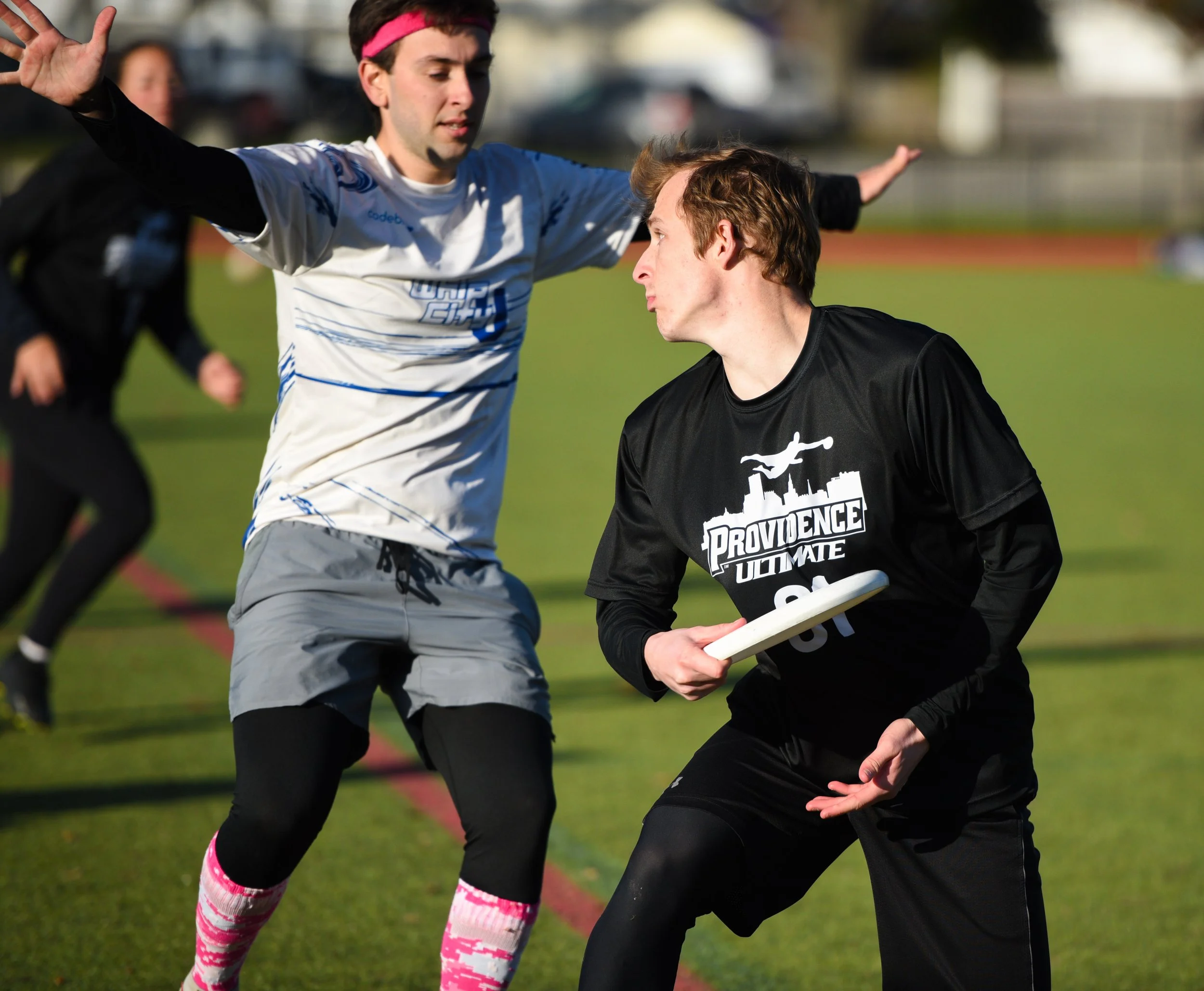 Two young men playing ultimate frisbee on a field, one wearing a gray and white jersey with a pink headband, and the other wearing a black jersey and holding a frisbee.