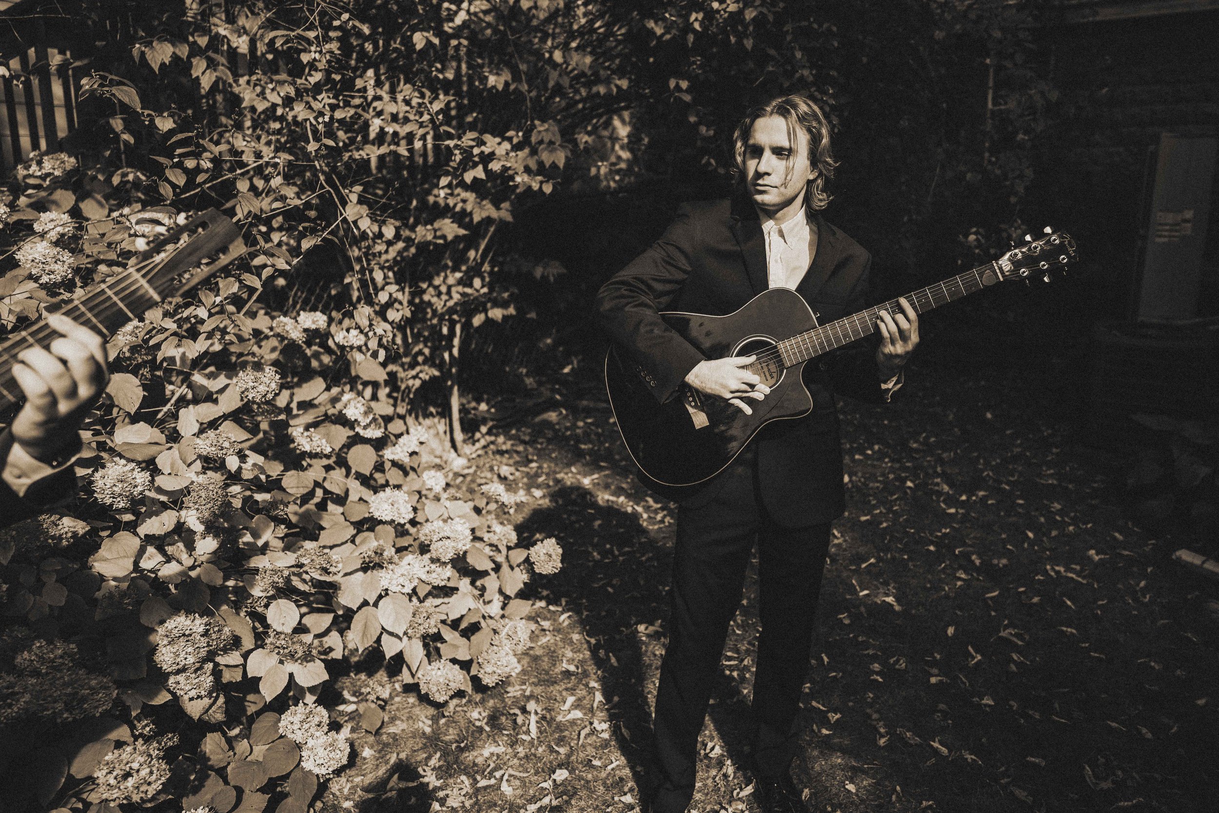 A young man in a suit playing an acoustic guitar outdoors at night, with bushes and leaves in the background.