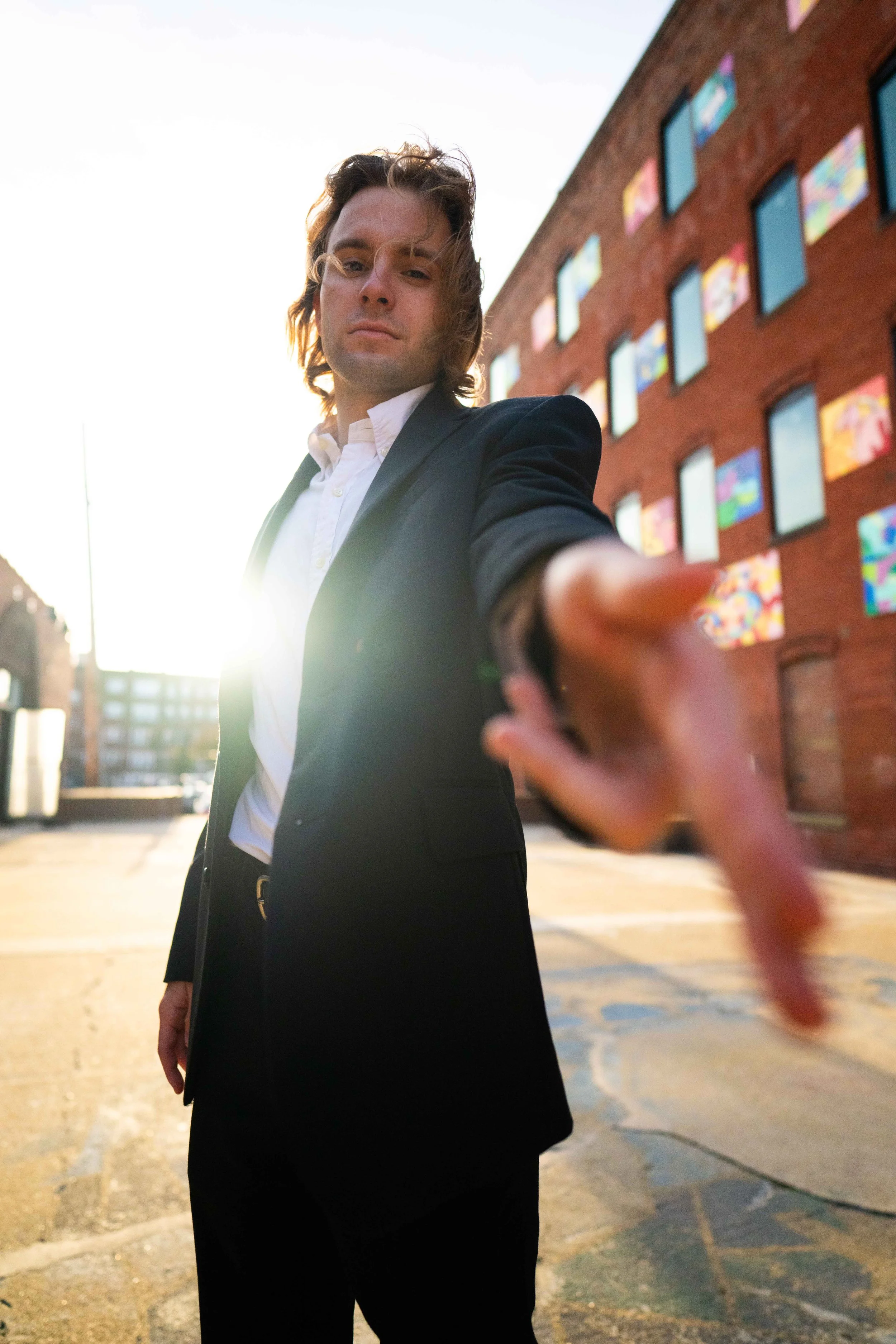 A man in a black suit and white shirt standing outdoors at sunset, extending his hand toward the camera with a city building and colorful windows behind him.