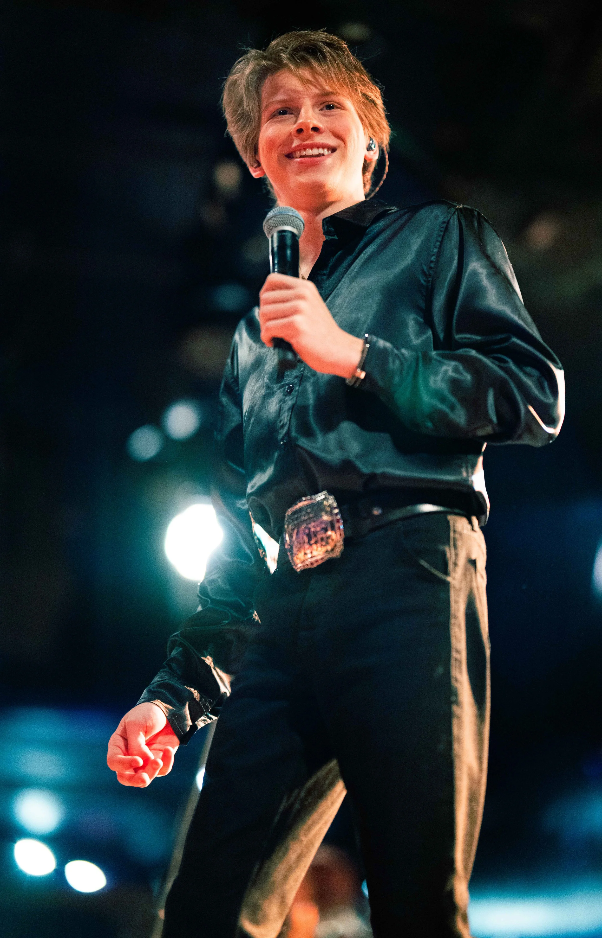 A young boy dressed as a police officer, holding a microphone, smiling on stage under bright lights.