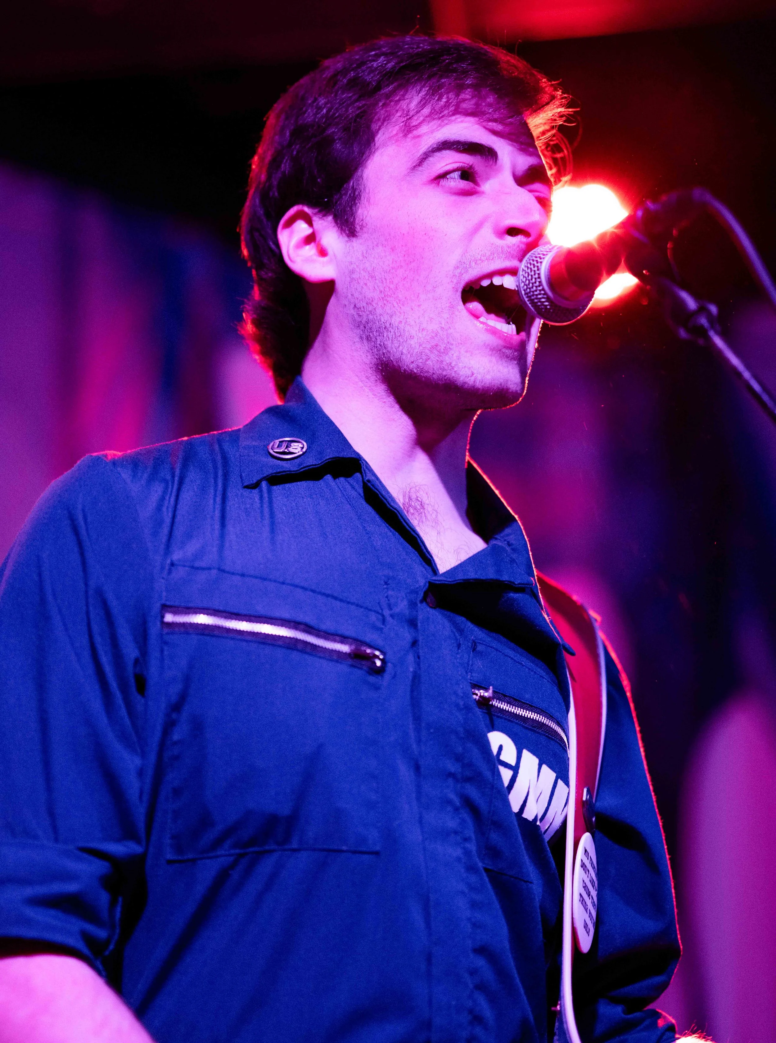 Young man singing or speaking into a microphone on stage, wearing a dark blue shirt with zippers, with colorful stage lighting in the background.