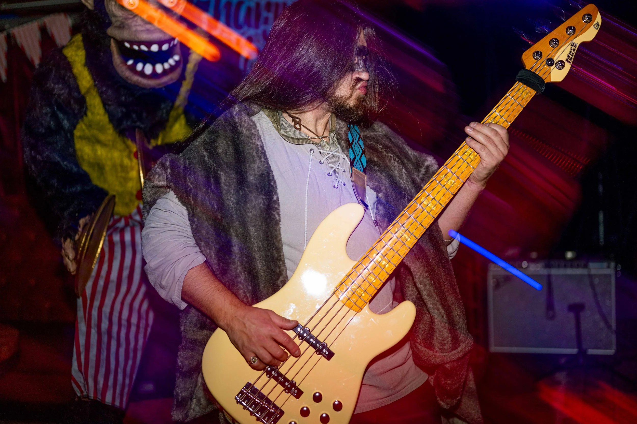 A man playing a yellow electric bass guitar on stage, with long hair and face paint, and a person in the background wearing a mask and colorful clown-like costume.