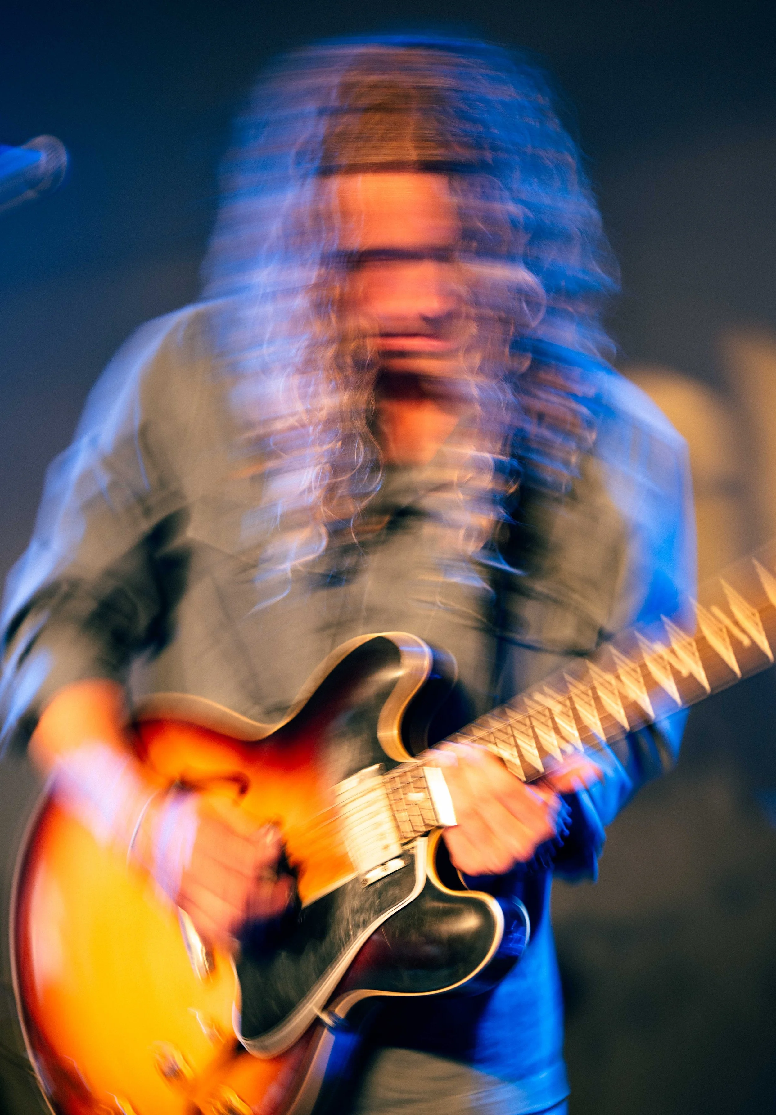 Blurred image of a musician playing an electric guitar on stage with colorful lighting.
