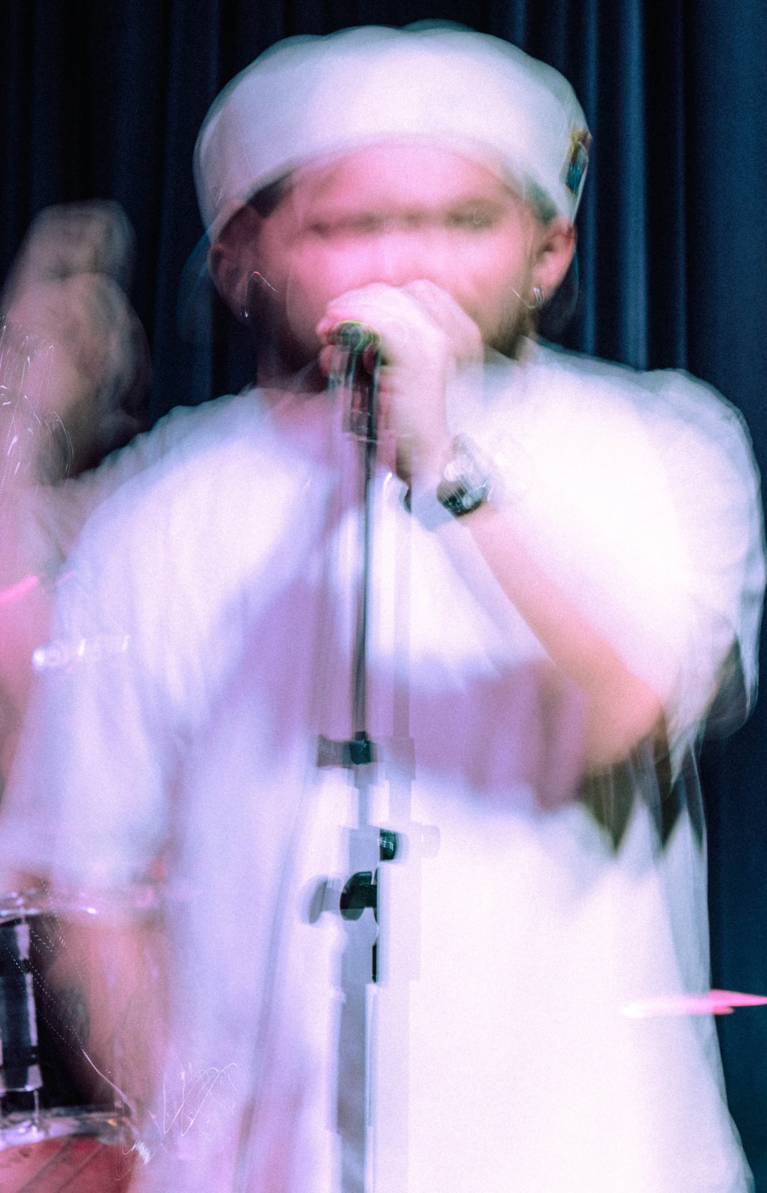 Blurry image of a person wearing a white shirt and a cap, speaking into a microphone with a dark background.