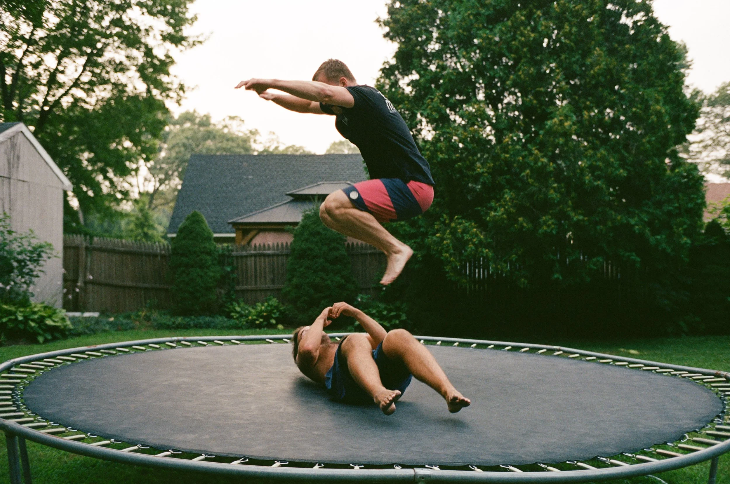 Two people are bouncing on a trampoline in a backyard, one is jumping into the air while the other is lying on the mat.