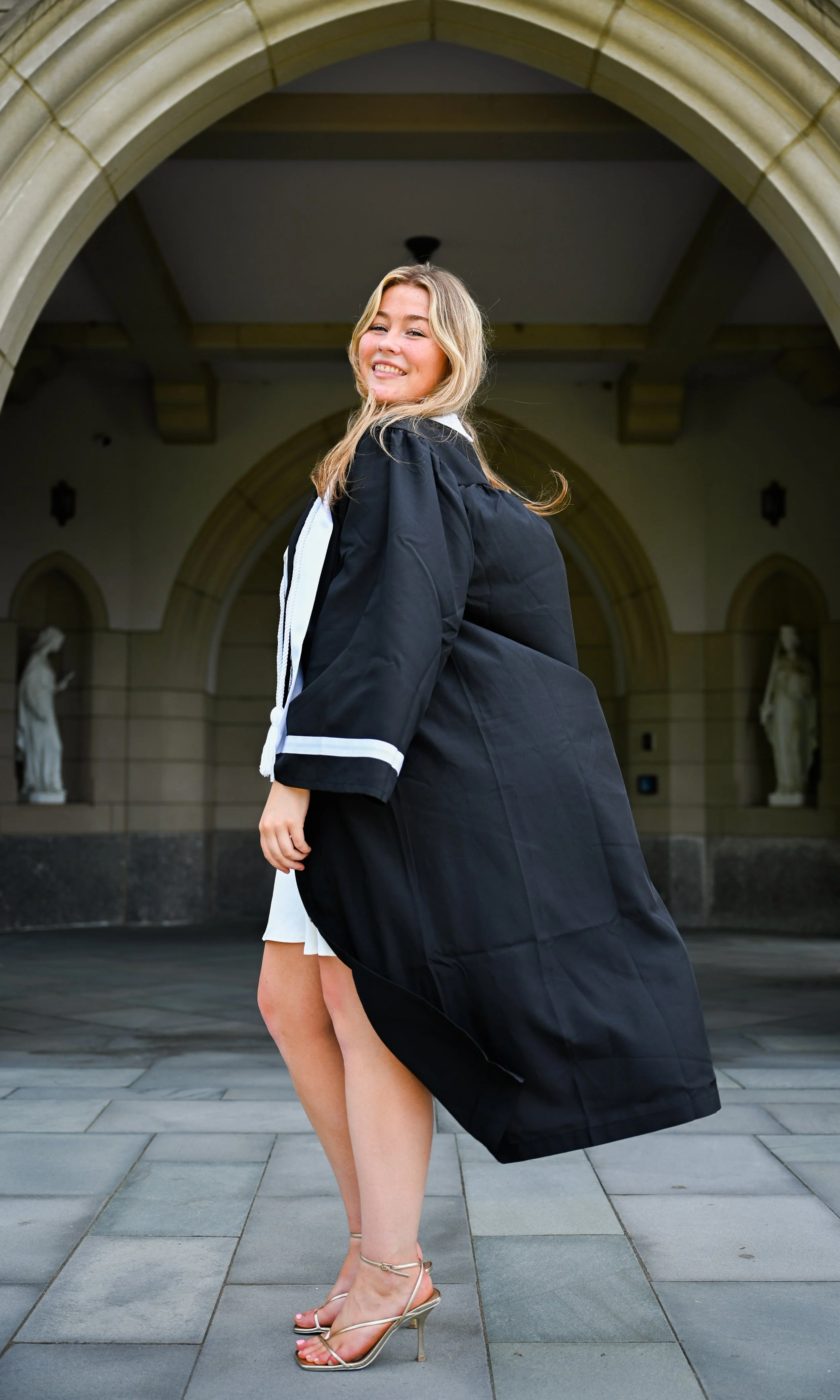 A woman in a graduation gown and heels standing outdoors in front of an arched building with statues.