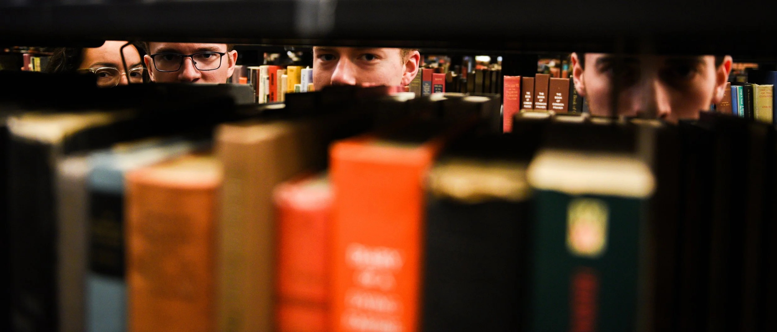 Three people peering through shelves of books in a library, with only their eyes and faces visible over the book spines.