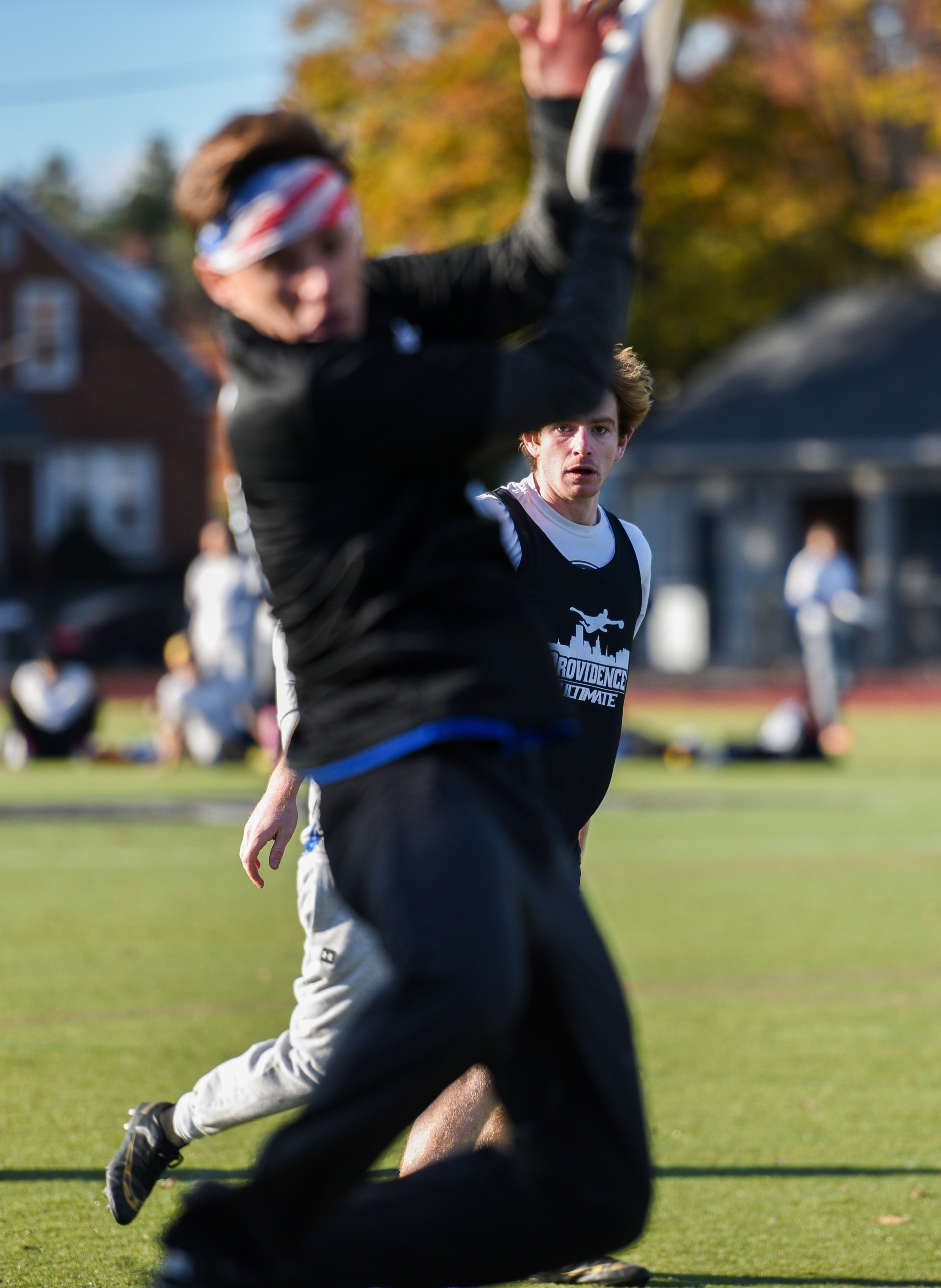 A person running on a track with other athletes and a man standing in the background, during daytime with fall foliage trees.