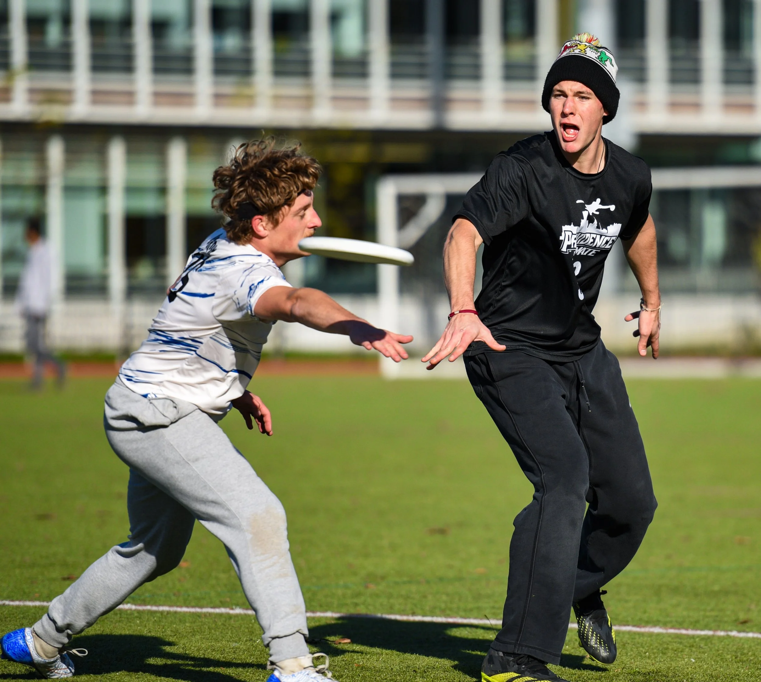 Two young men playing frisbee on a football field with a modern building in the background. One man is throwing the frisbee while the other is reaching to catch it.