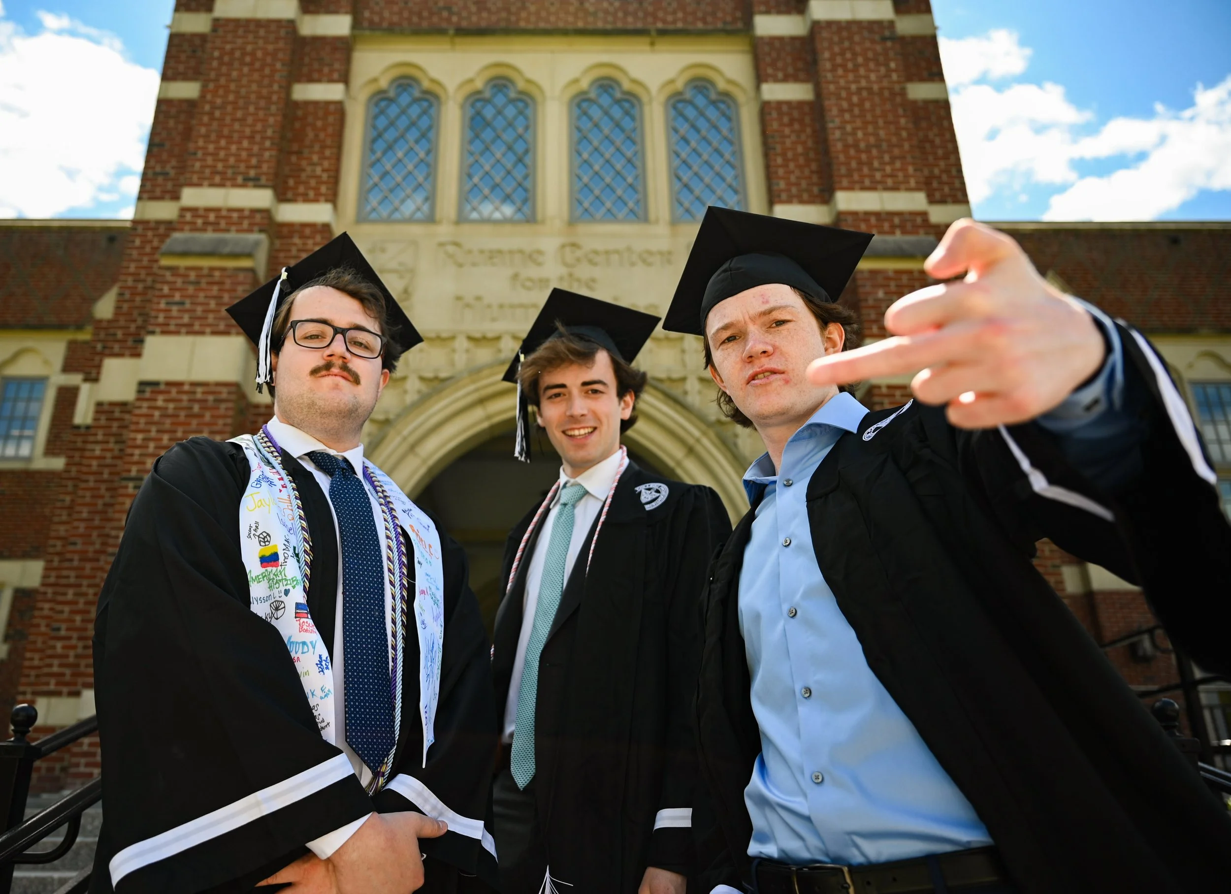 Three young men in graduation caps and gowns standing in front of a university building, with one pointing towards the camera and smiling.