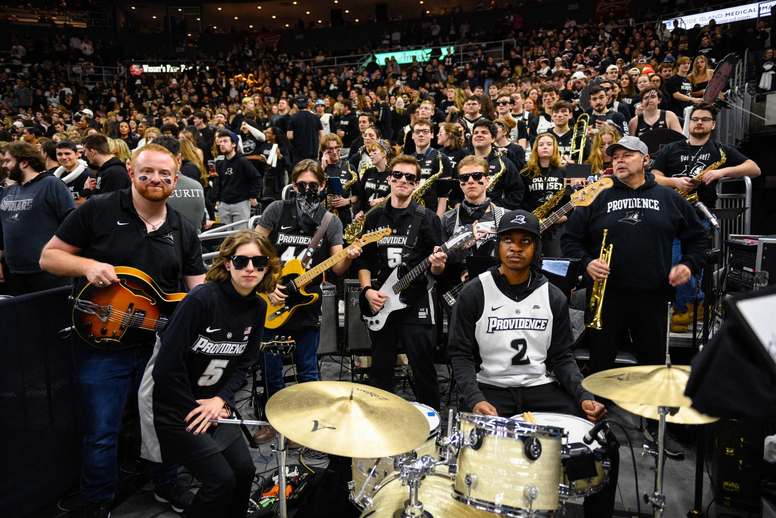 Group of young musicians in athletic jerseys and casual clothing, posing with their musical instruments at a sports arena filled with spectators.