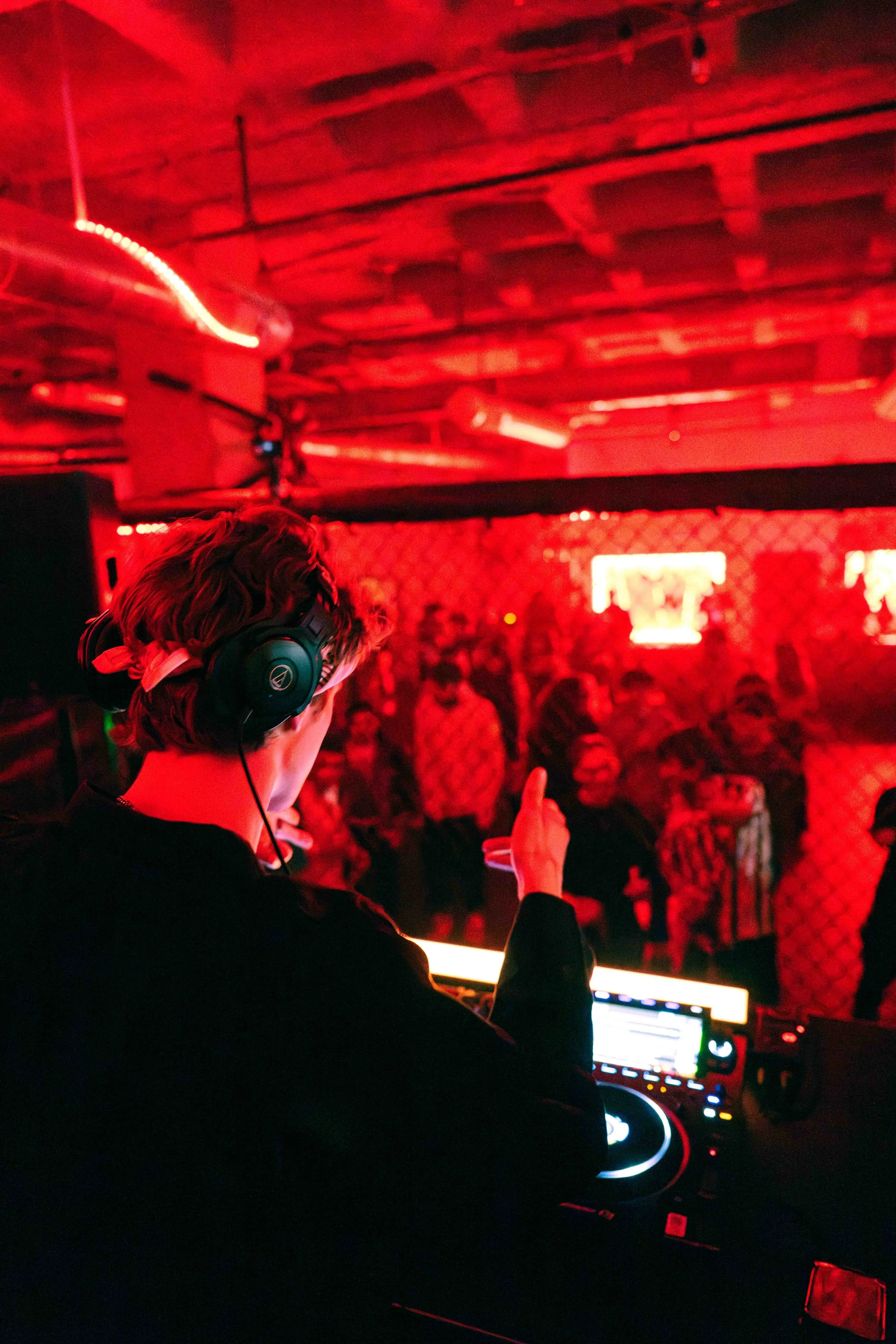 A DJ with headphones performing at a nightclub or music event, with a crowd of people dancing in the background illuminated by red lighting.