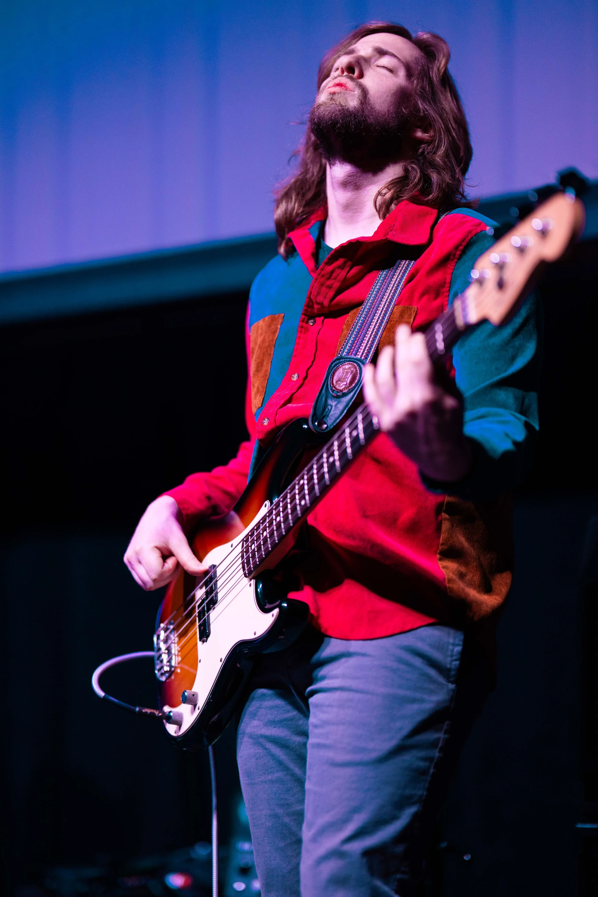 Man with long hair and beard playing electric guitar, eyes closed, wearing colorful patchwork shirt, standing on stage.