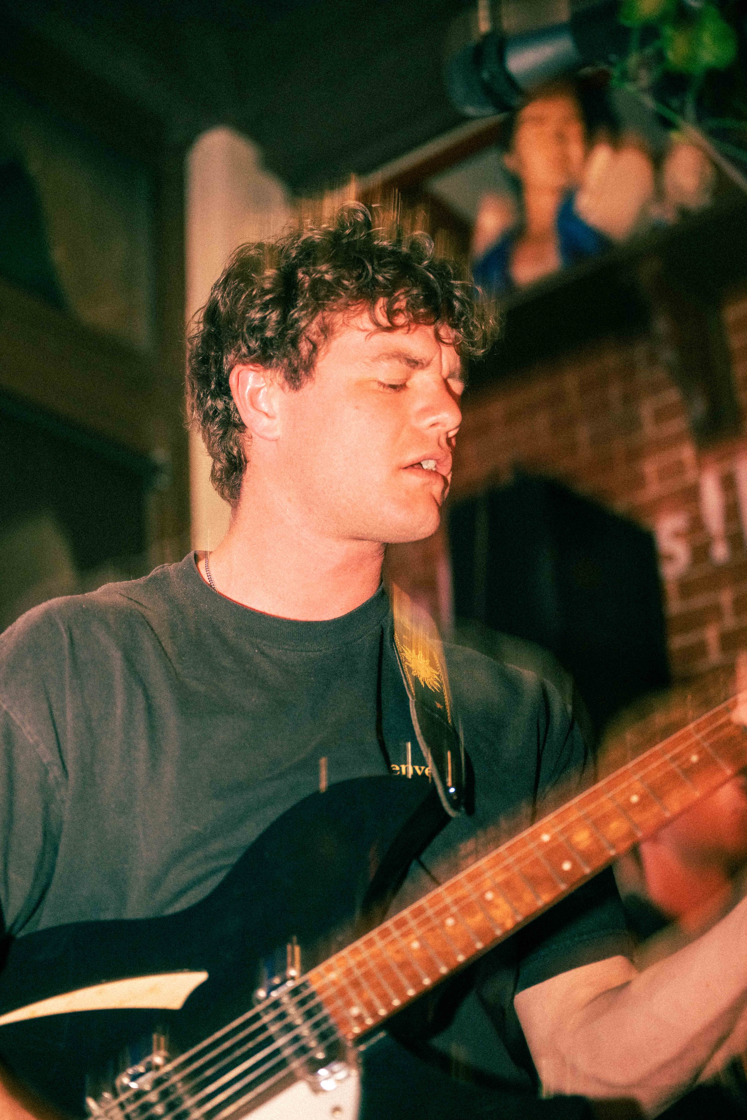 A man with curly hair playing an electric guitar in a dimly lit room, with a portrait or painting of someone hanging on a brick wall in the background.