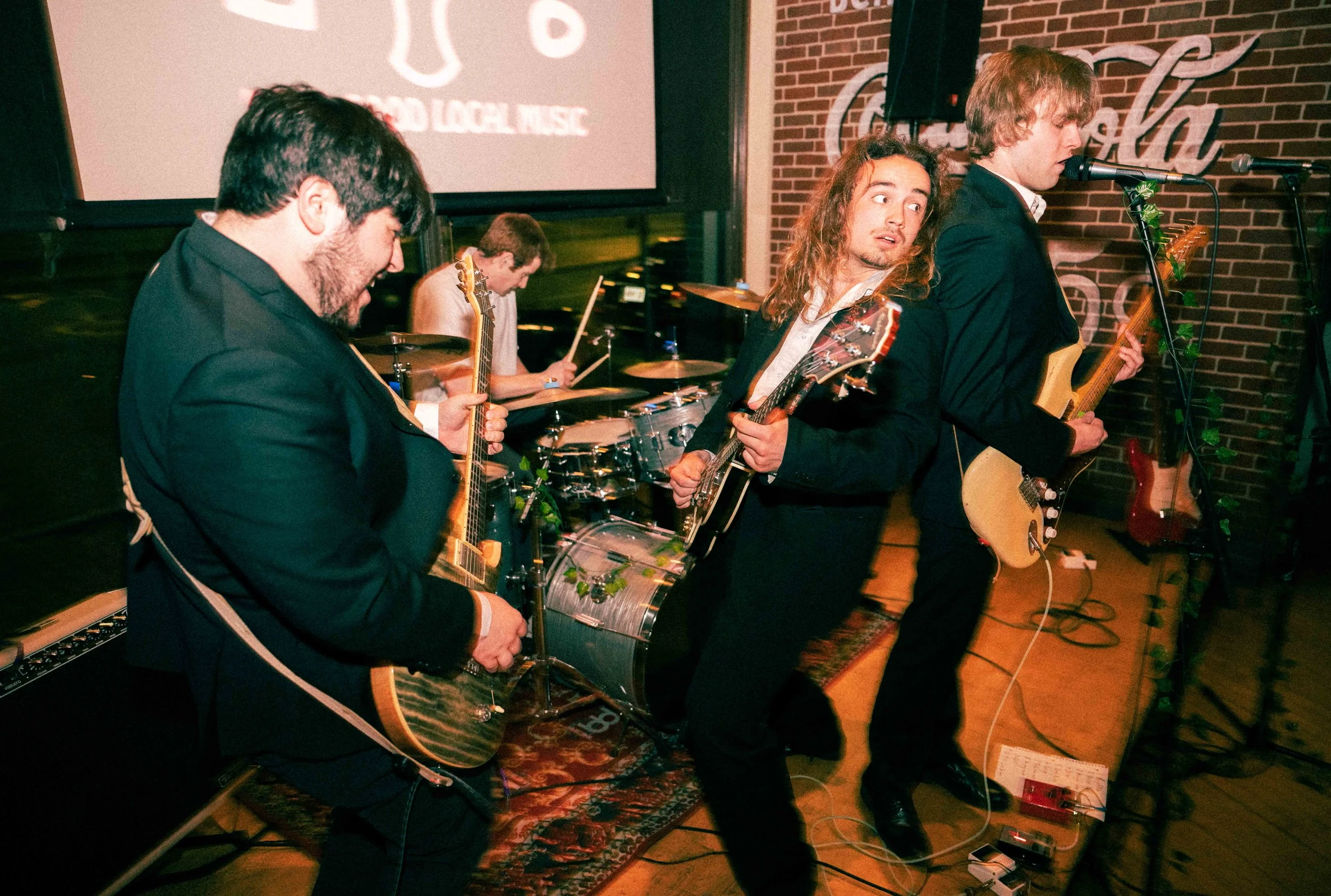 A four-member band performing on stage with guitars and drum set, with a backdrop featuring a logo and brick wall.