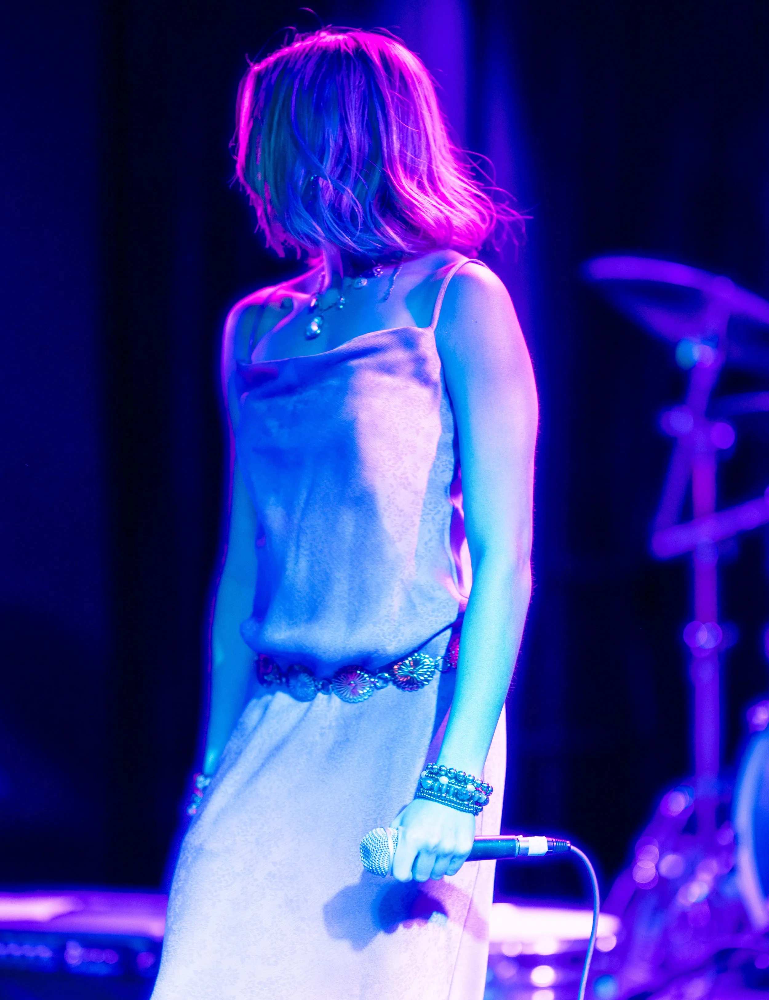 Woman with colorful hair performing on stage, holding a microphone, illuminated by vibrant purple and blue lighting.