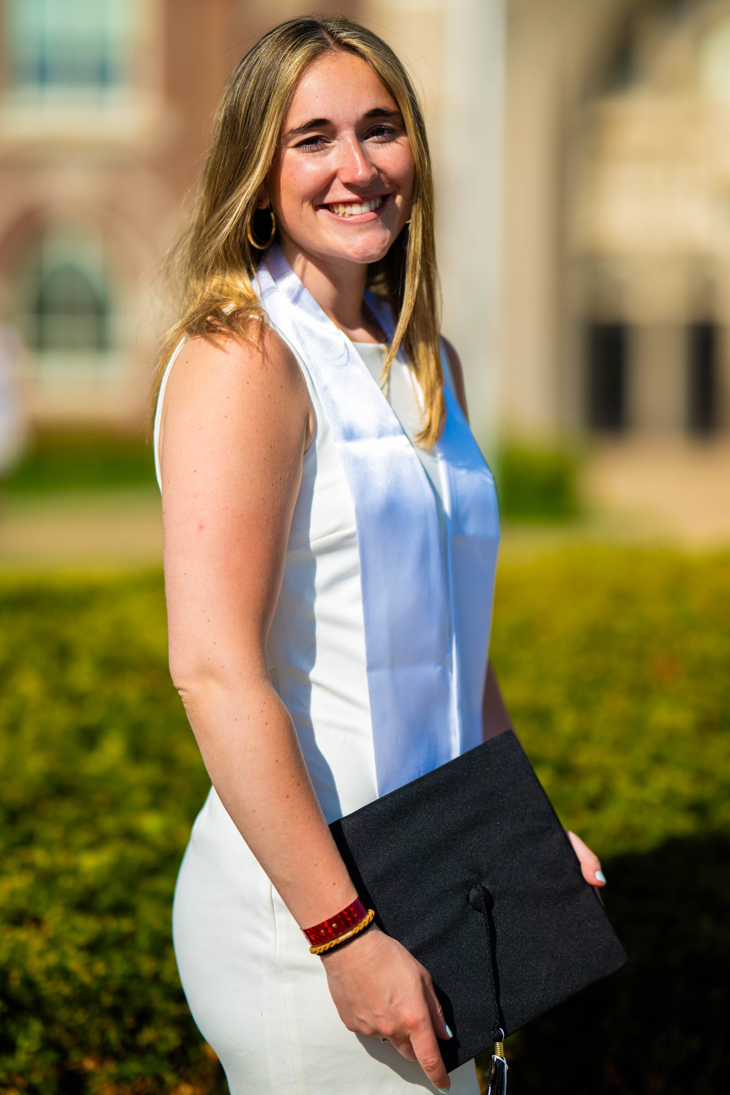 A young woman in a white dress holding a graduation cap, smiling outdoors with a building in the background.
