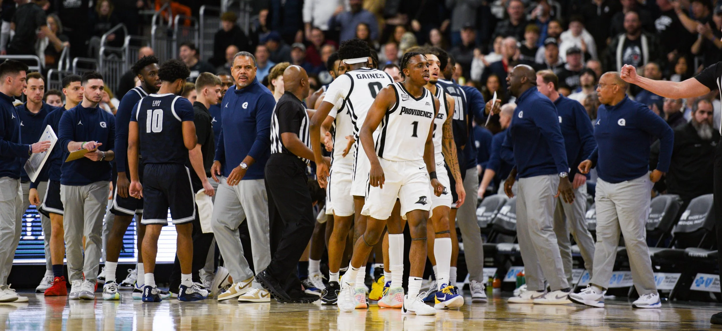 Basketball players and coaches on a court during a game timeout, some standing and some sitting, with a crowd in the background.