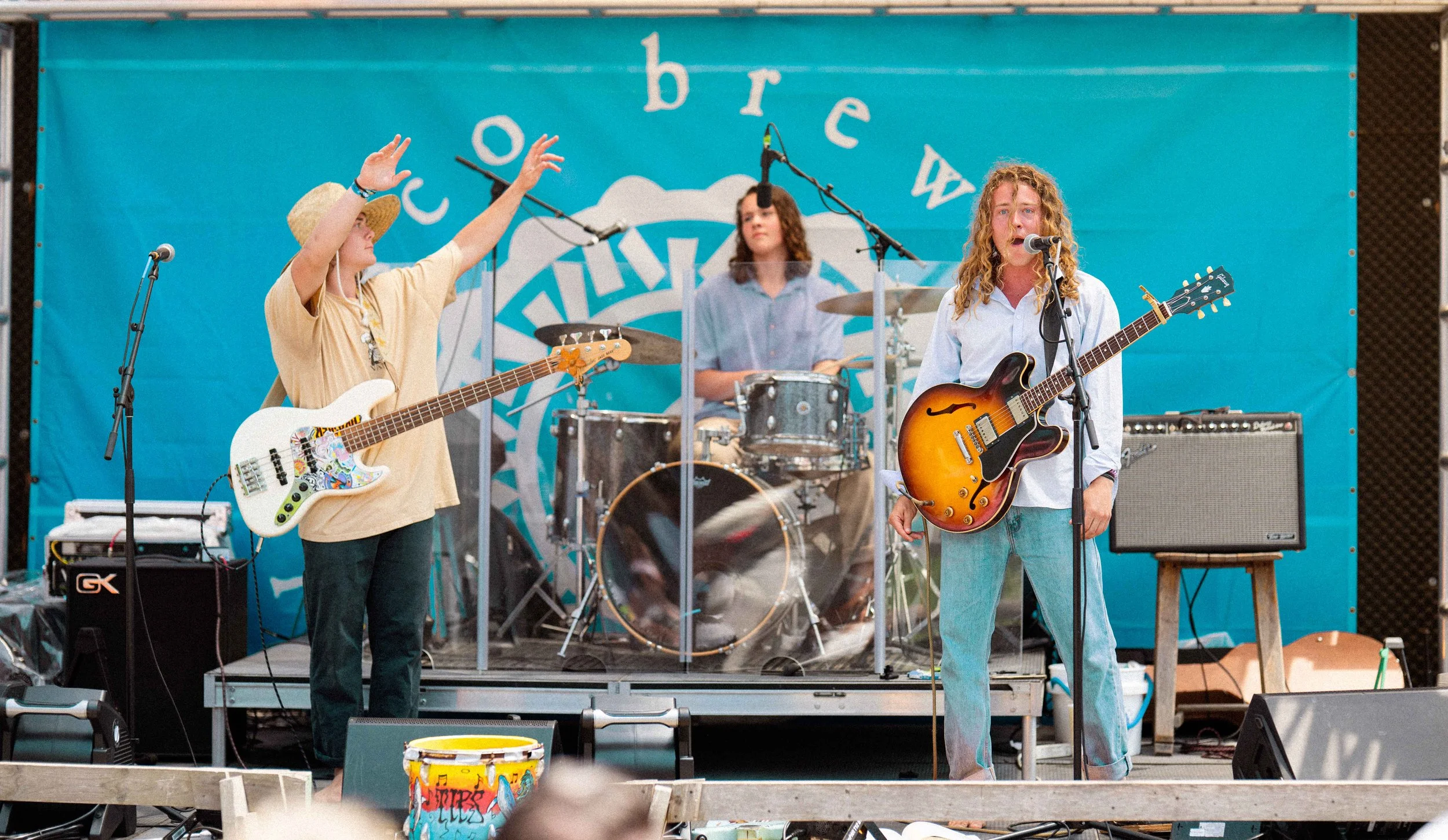 A band performing on a stage with a blue backdrop that has the words 'cow breast' on it. The band includes a woman with curly hair singing into a microphone and playing an electric guitar, a woman with long hair playing drums, and another woman weari