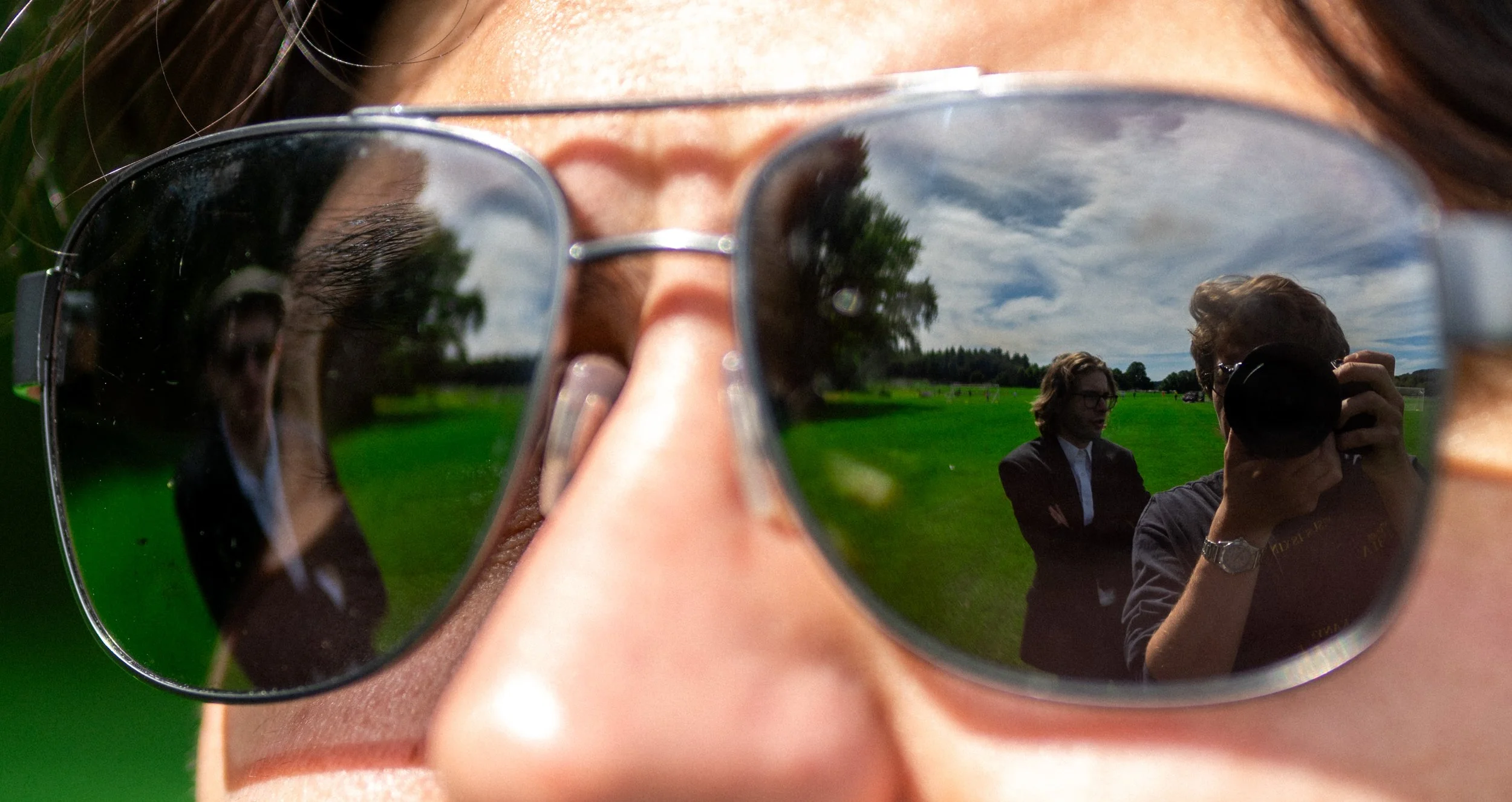 Close-up of person wearing sunglasses taking a reflection photo on the sunglasses' lenses, showing a grassy field and people in the background.