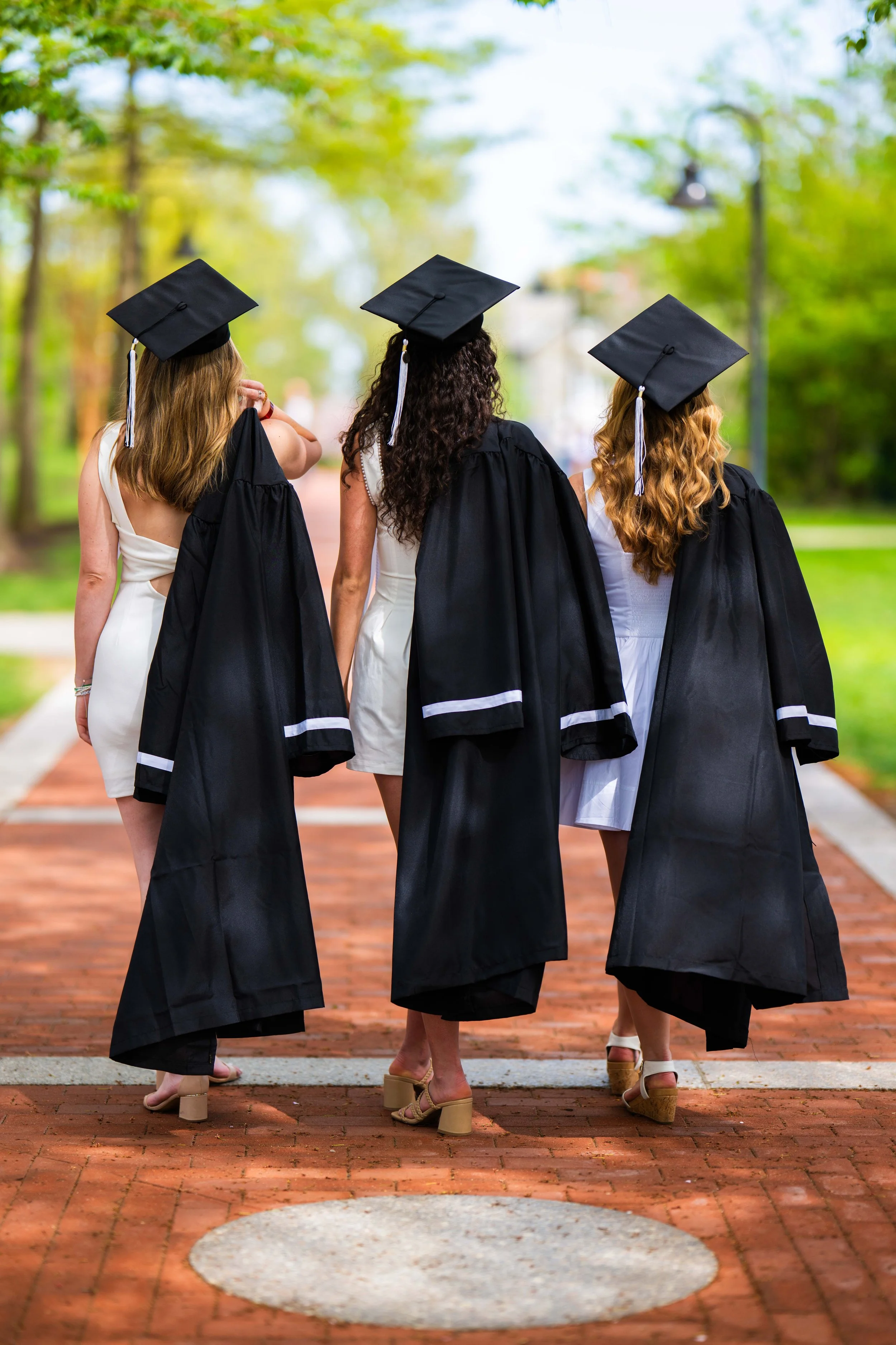 Three women in black graduation gowns and caps walking on a brick path outdoors, with green trees in the background.