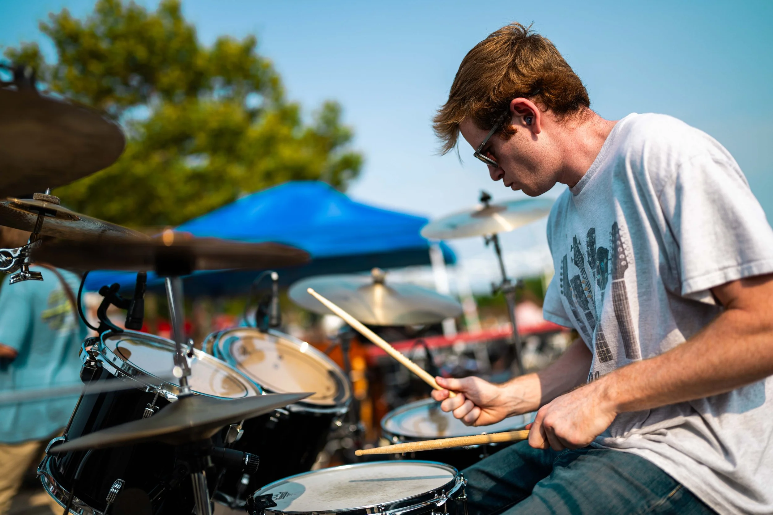 A young man playing the drums outdoors during daytime, wearing a gray t-shirt and sunglasses, with a blue canopy and trees in the background.