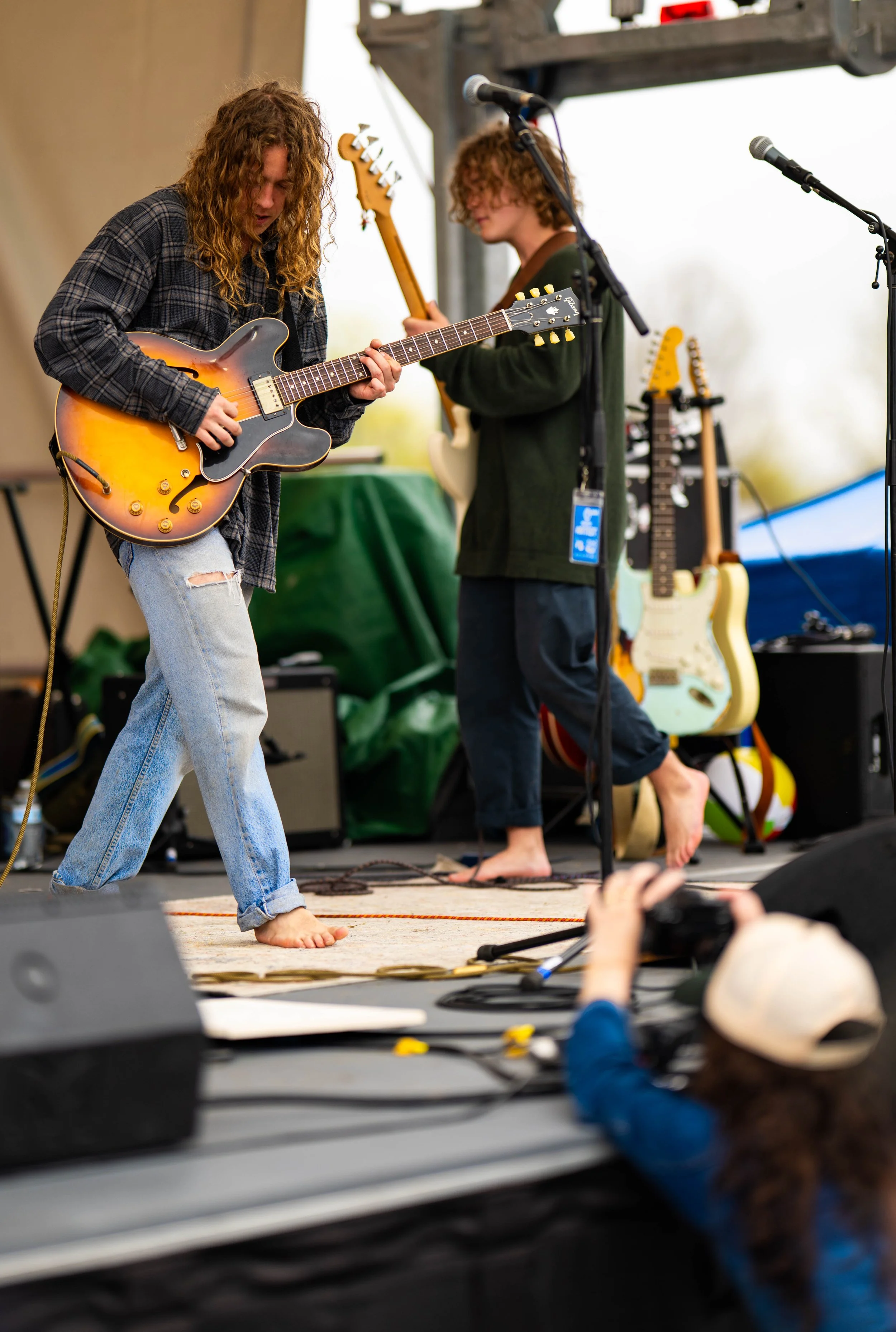 Two young men with curly hair playing guitars on a stage during a live outdoor music performance, with various guitars and equipment in the background.