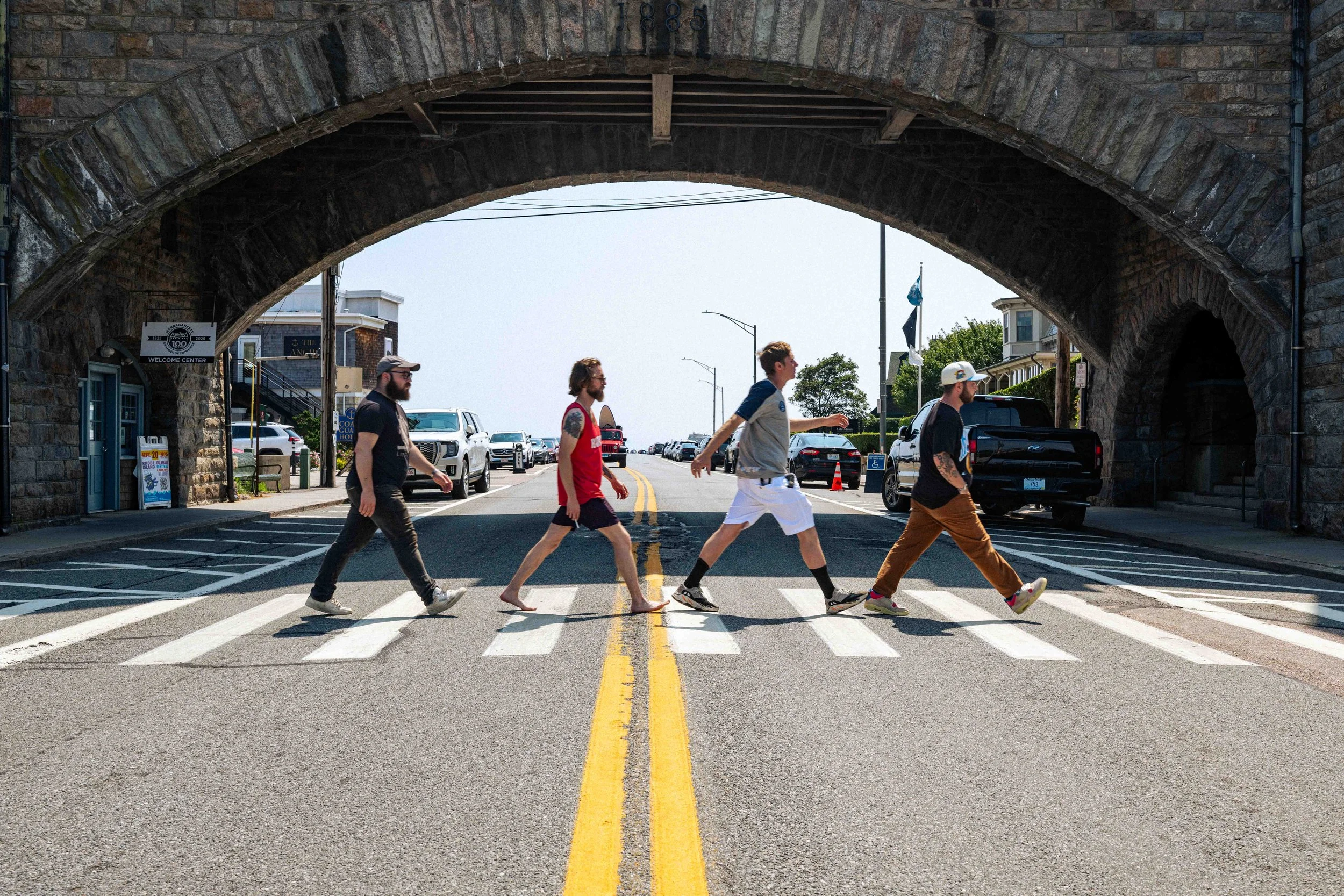 Four people walking across a pedestrian crosswalk under a stone bridge in an urban area with parked cars and buildings.