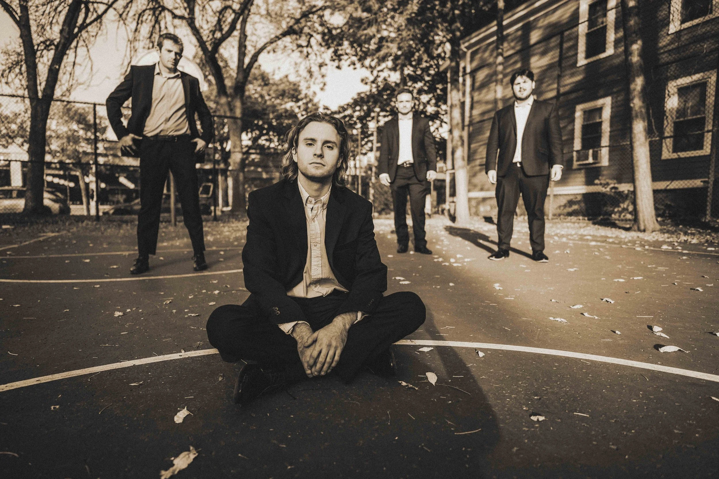 A group of five young men in suits pose on an outdoor basketball court in a suburban neighborhood during autumn. One man is sitting cross-legged on the ground in the foreground, while the other four stand behind him with serious expressions. The scen