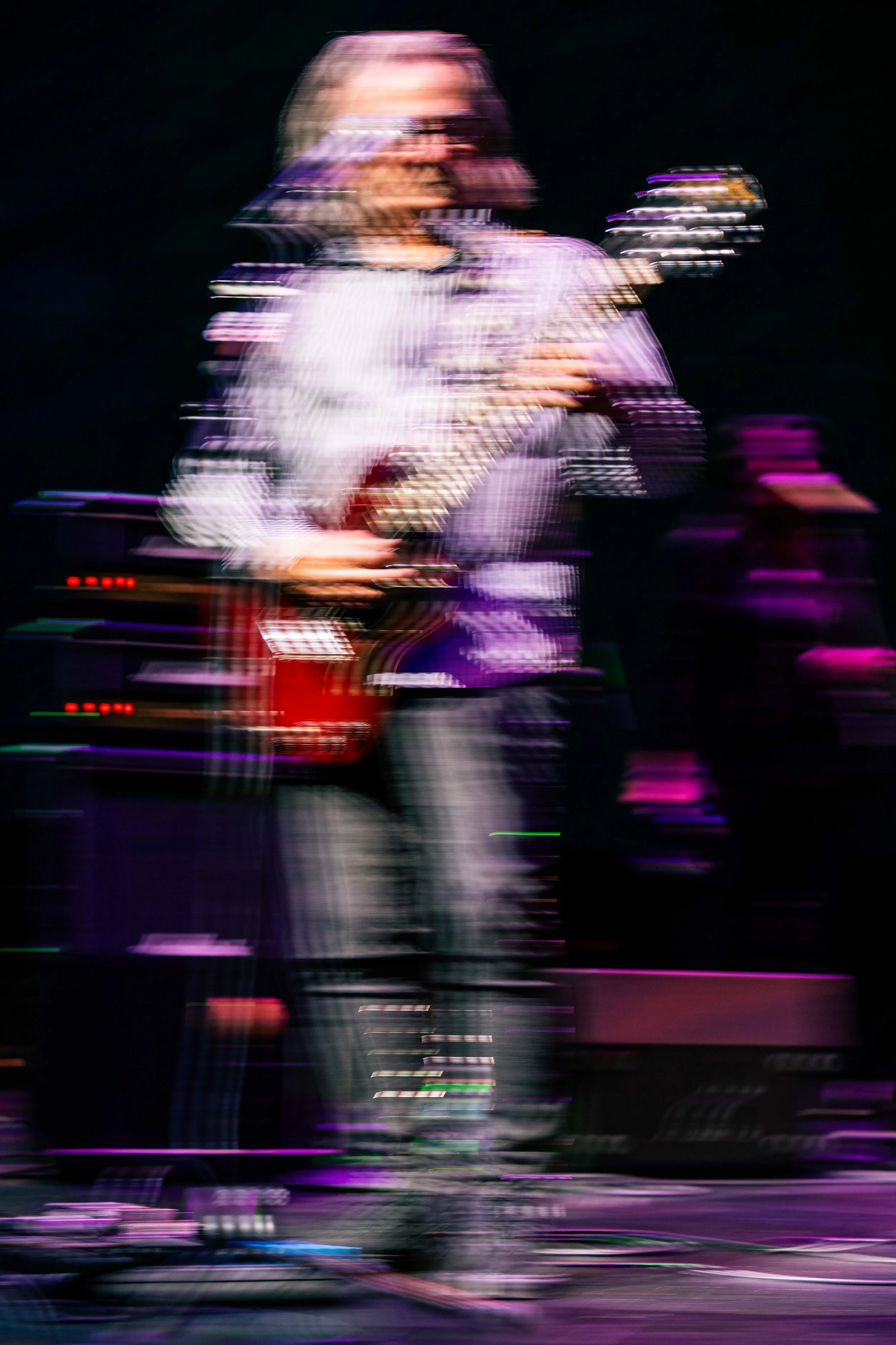 A person with light-colored hair playing an electric guitar on stage, captured with camera motion blur creating colorful streaks of light against a dark background.