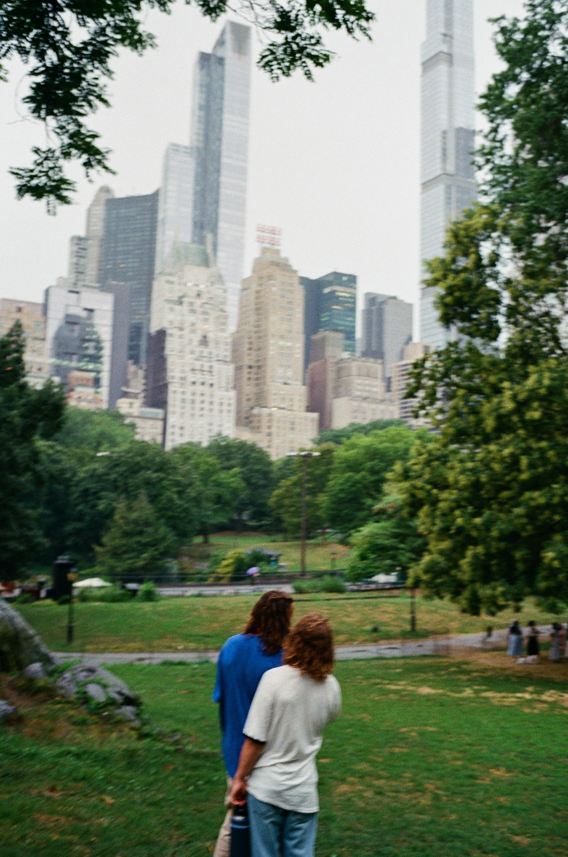Two men with brown hair, one in a white shirt and the other in a blue jacket, standing in a green park with tall buildings in the background, during an overcast day.