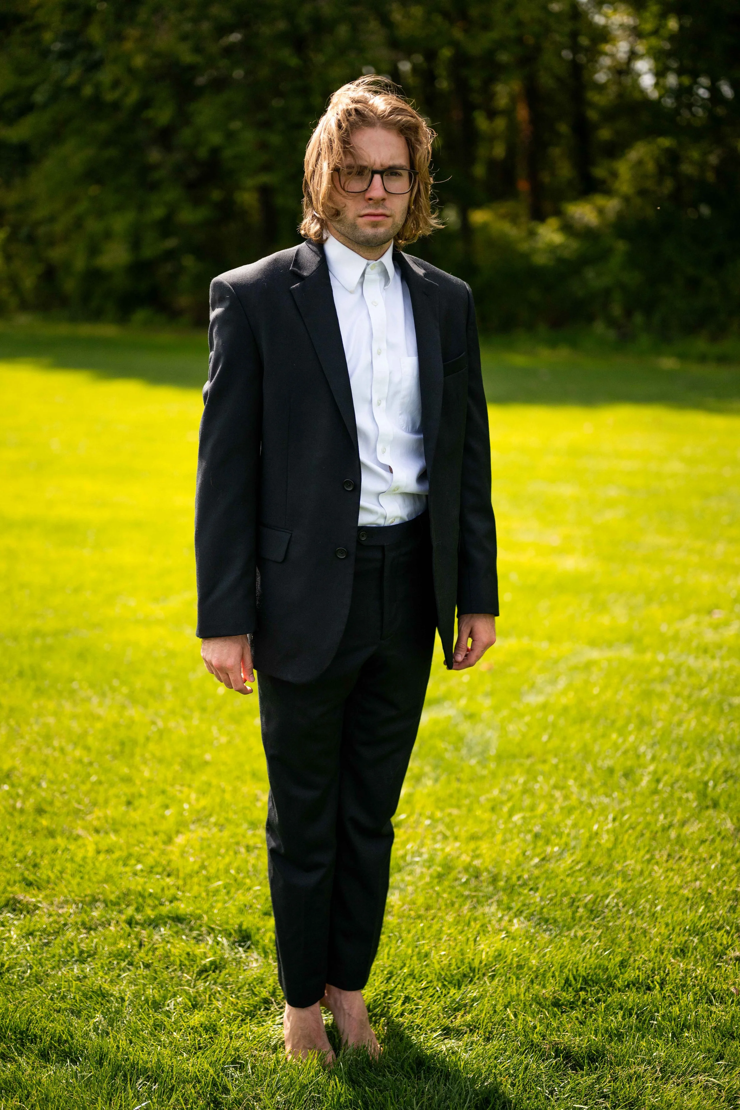 Man in a black suit and glasses standing barefoot on a grassy field with trees in the background, looking serious.