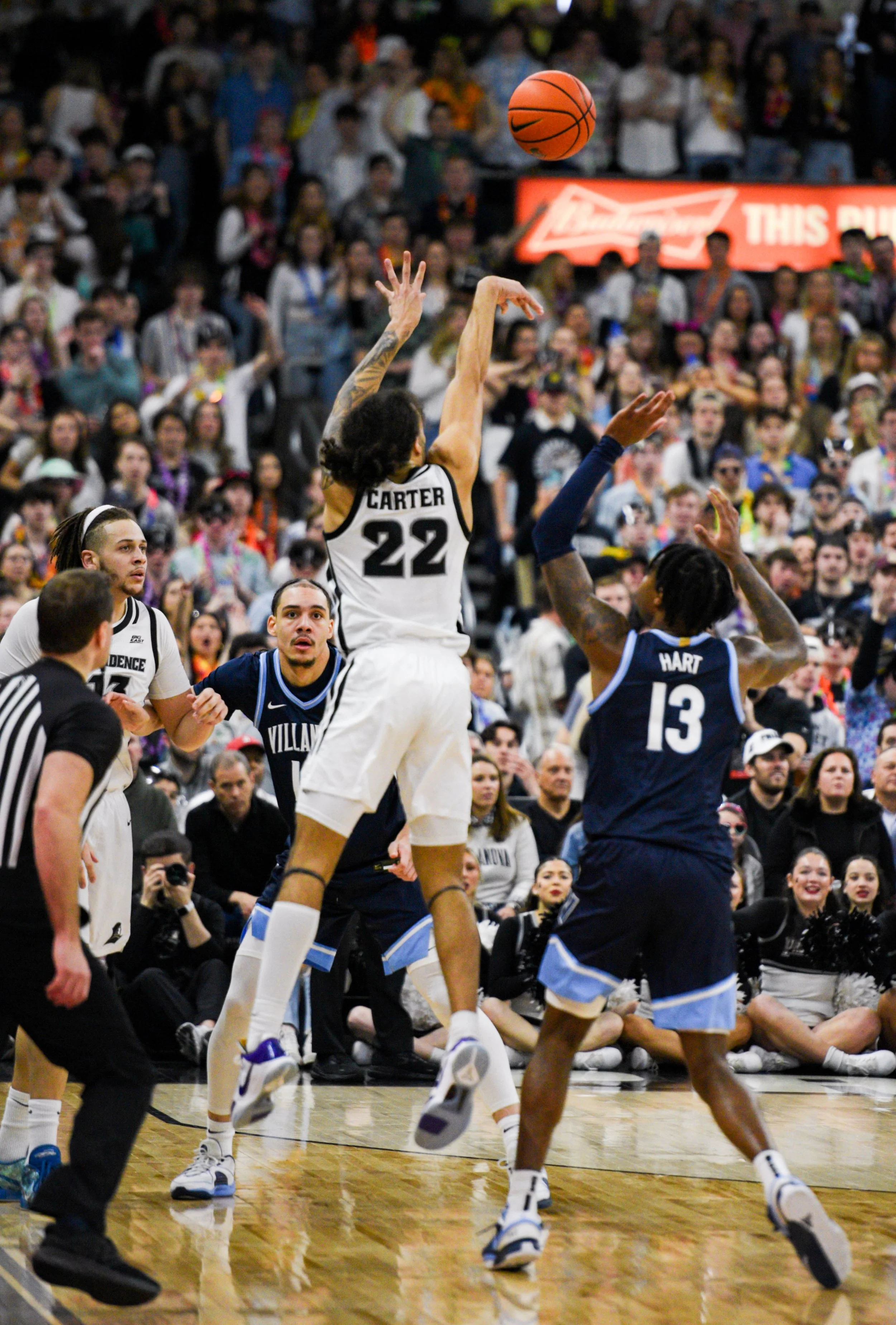 A basketball game with players in white and navy jerseys. Player #22 from the white team jumps to shoot or pass, while player #13 from the navy team raises his arms to block. The crowd in the background is watching intently.