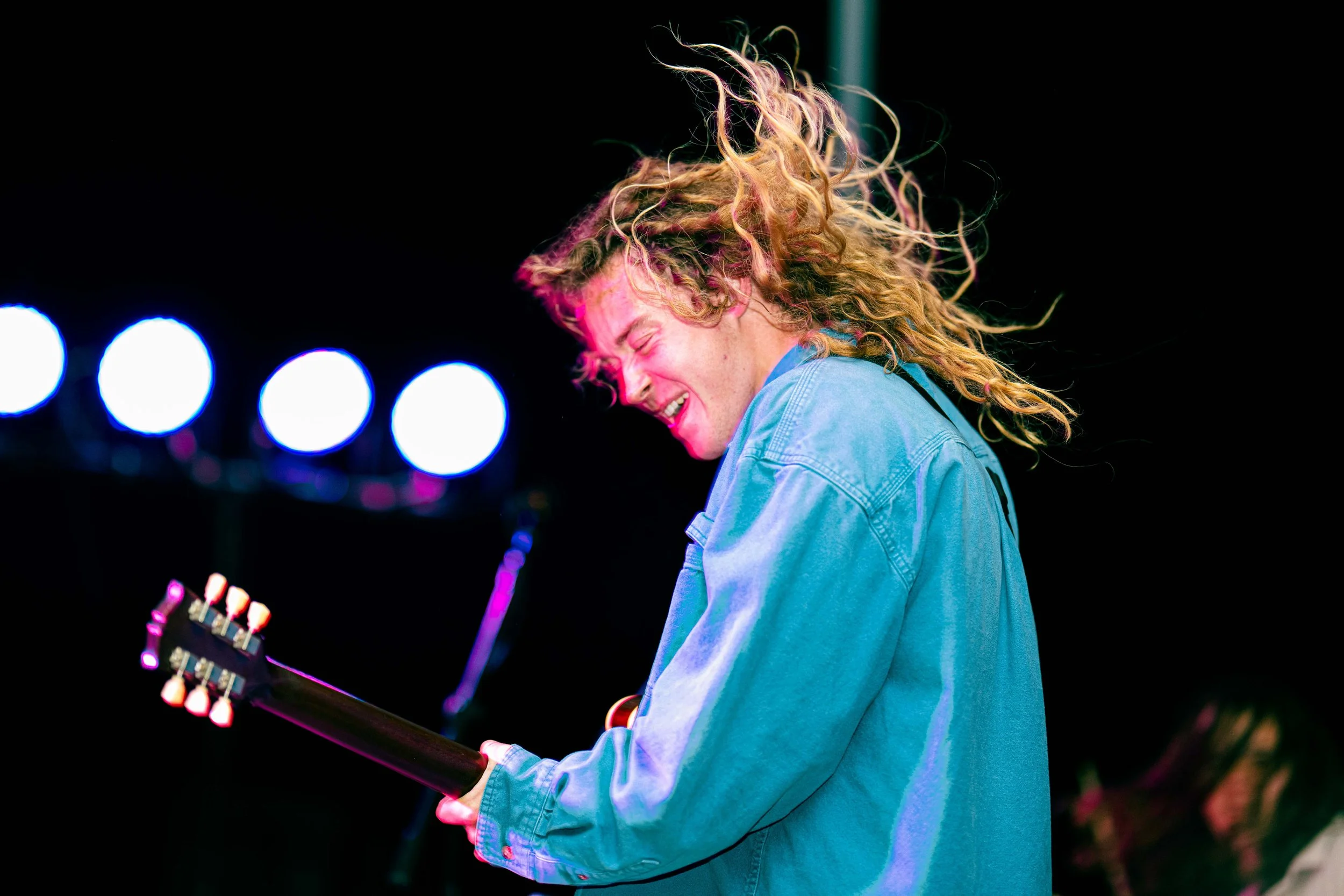 A young man with long, curly blonde hair playing an electric guitar on stage at a live music event, smiling and enjoying the moment with bright stage lights behind him.