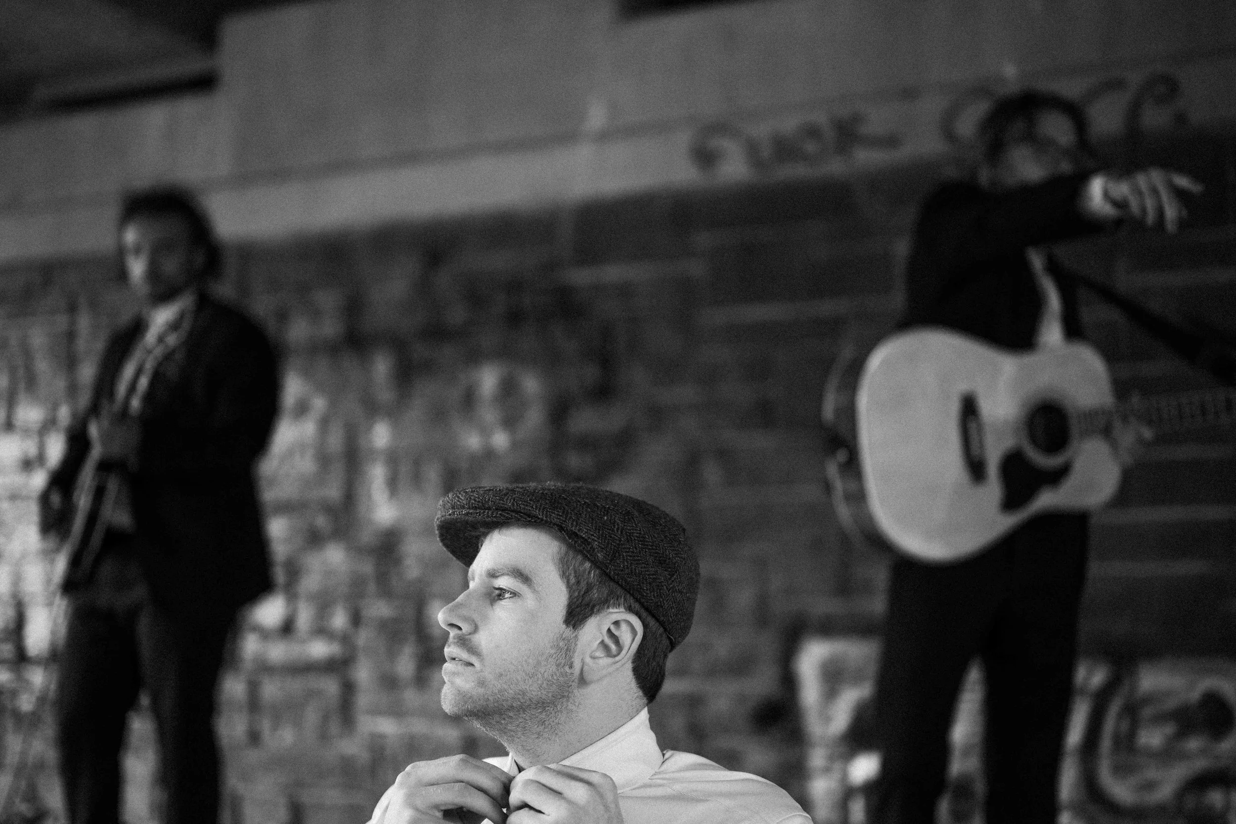 A black and white photo shows a young man in the foreground adjusting his collar, wearing a flat cap and a white shirt. In the background, two musicians are playing guitars, one with a microphone, against a brick wall.