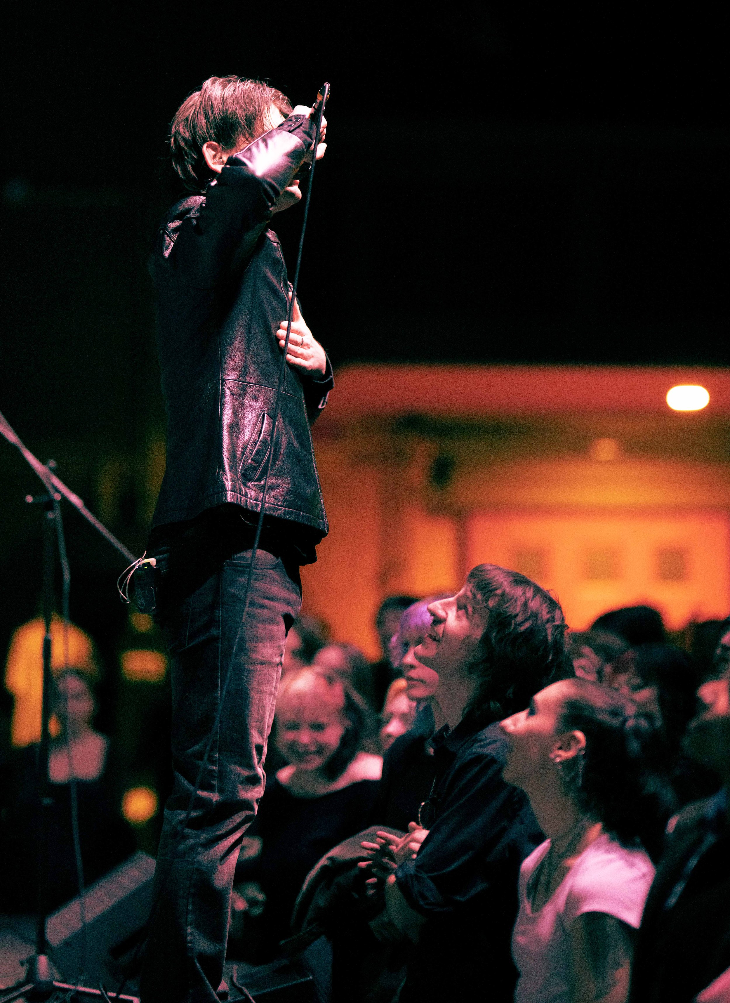 Person on stage holding a microphone near their face, engaging with audience members seated in front, some smiling and looking up at them.