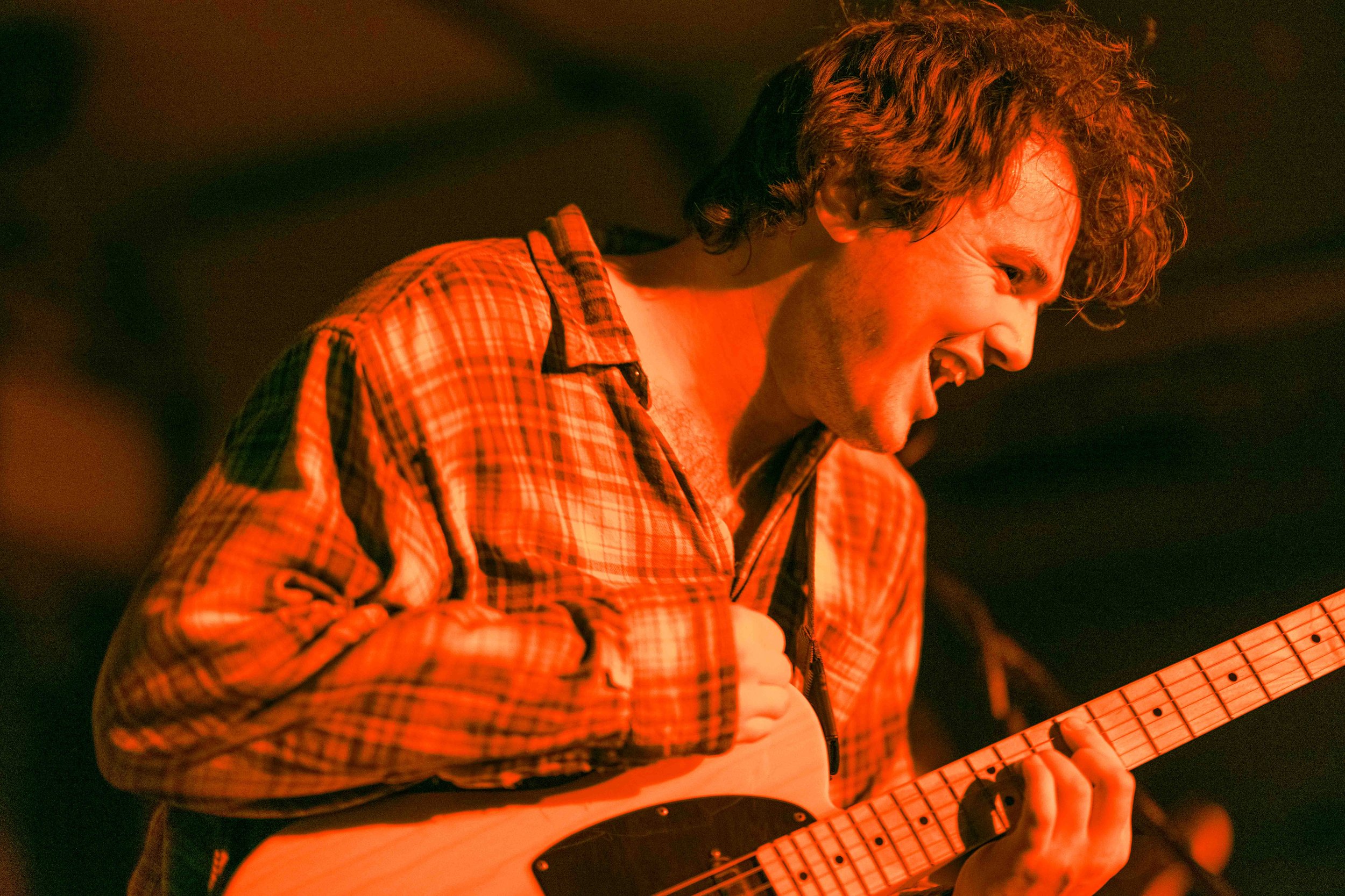 A man with curly hair wearing a plaid shirt passionately playing an electric guitar in a performance setting.