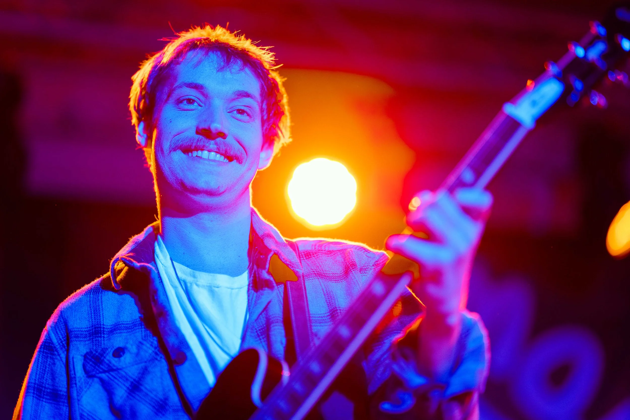 A young man with a smile playing an electric guitar on stage, illuminated by vibrant red, orange, blue, and purple stage lights.