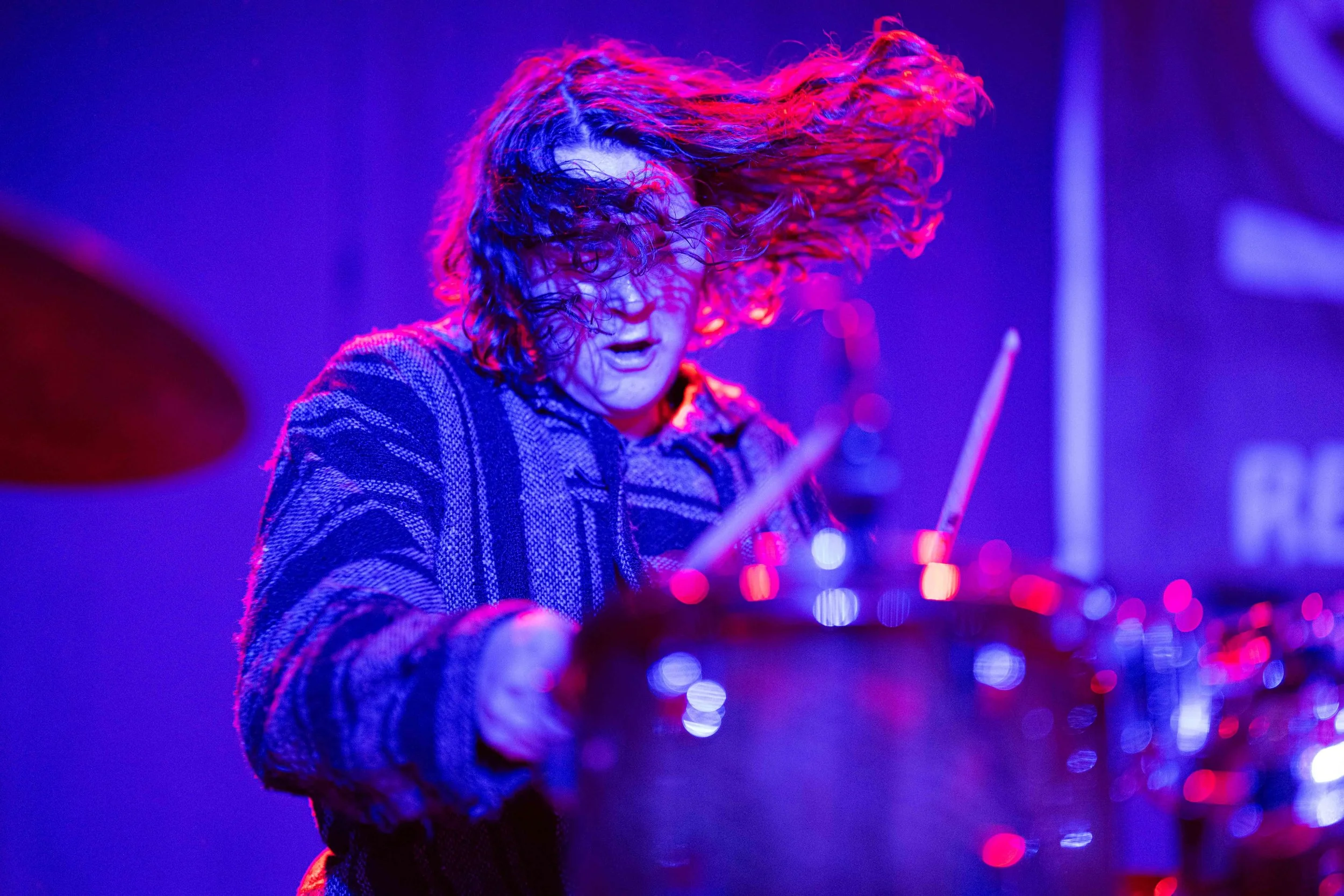 A musician with curly hair performing on stage with colorful lighting, intensely focused while playing the drums.