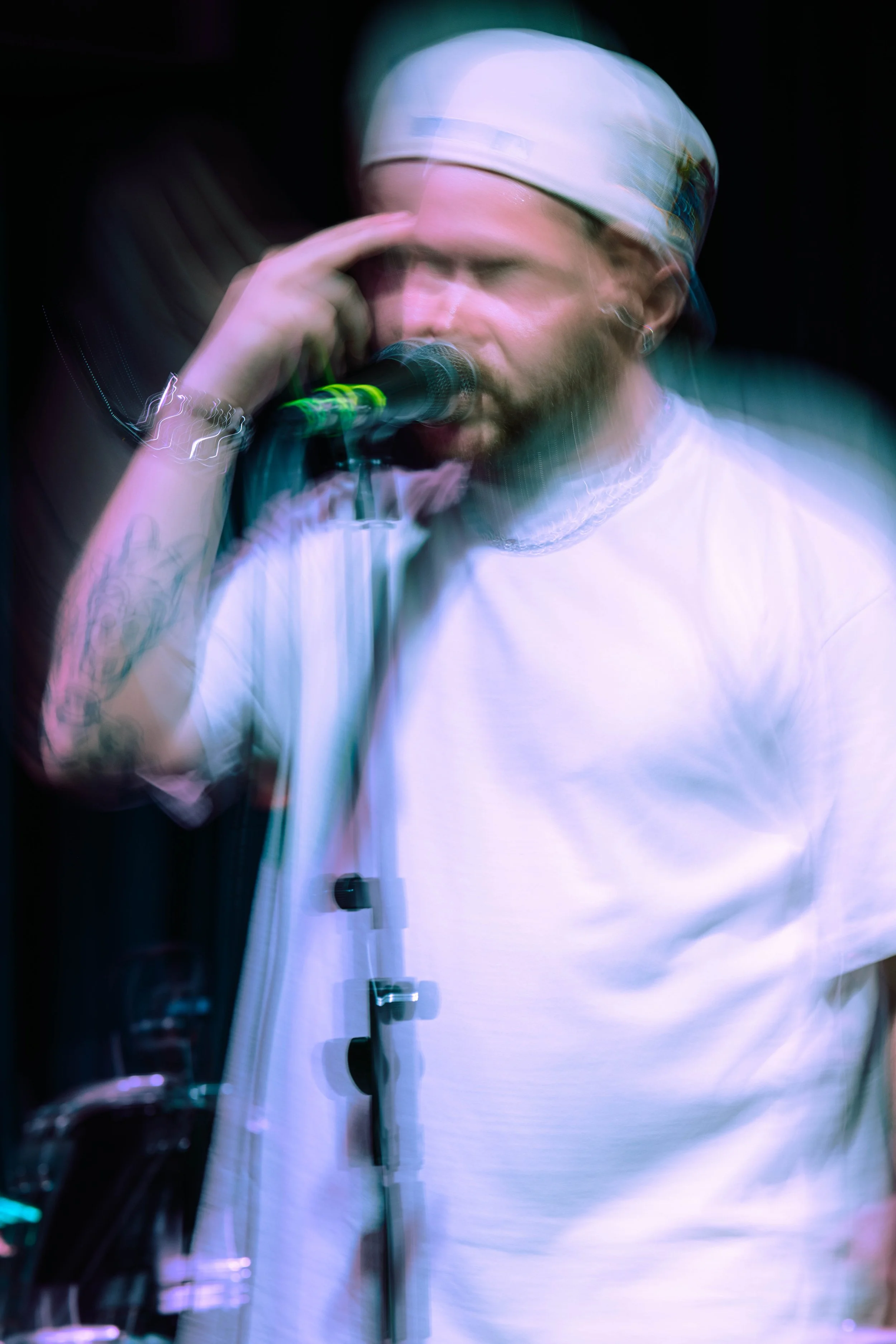 A man wearing a white chef's hat and a white shirt, standing in front of a microphone with his right hand touching the side of his forehead, while slightly leaning towards the microphone.
