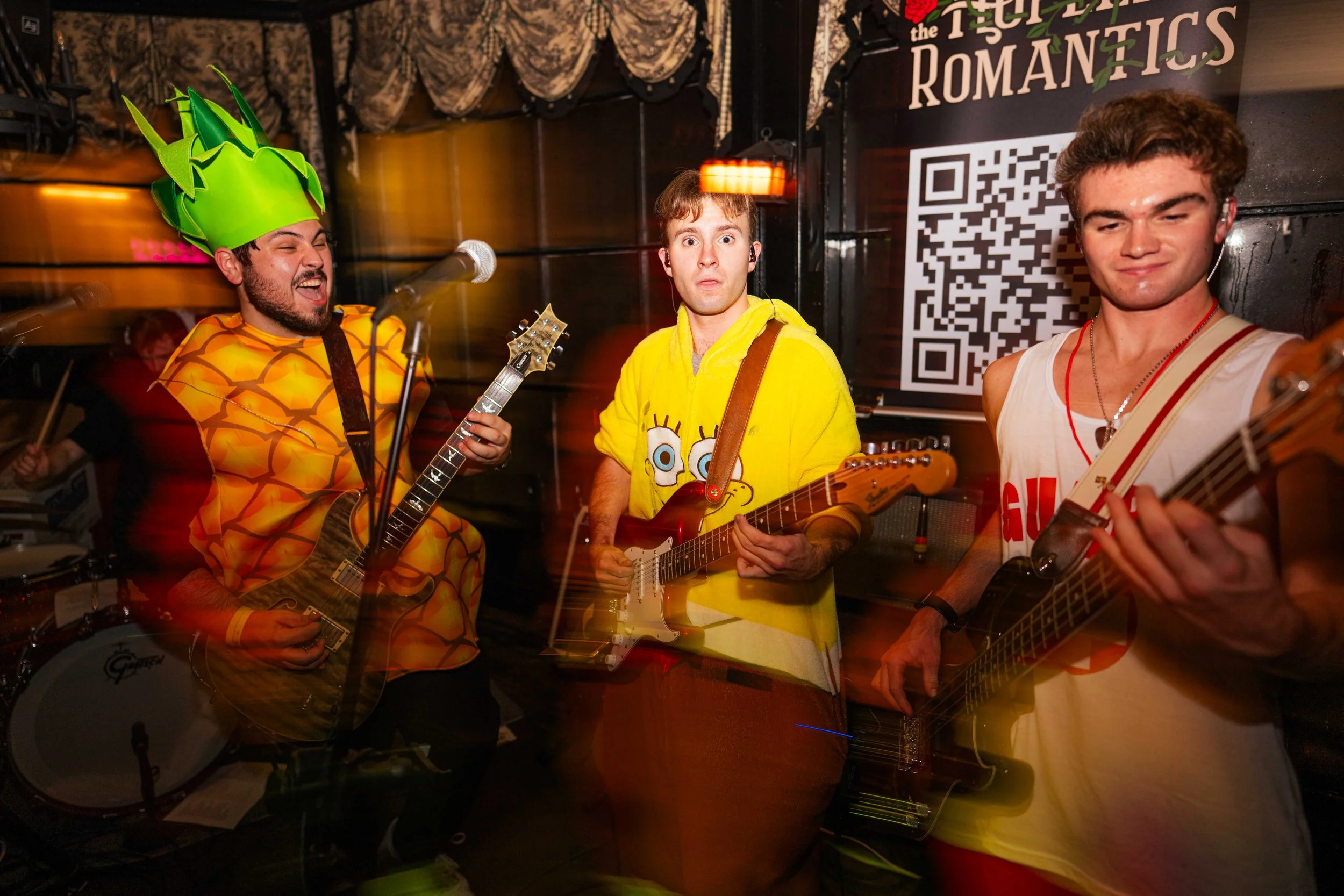 Three young men playing guitars and singing in a band, two wearing colorful costumes and one in a sleeveless shirt, in a dimly lit room with a sign and a QR code in the background.