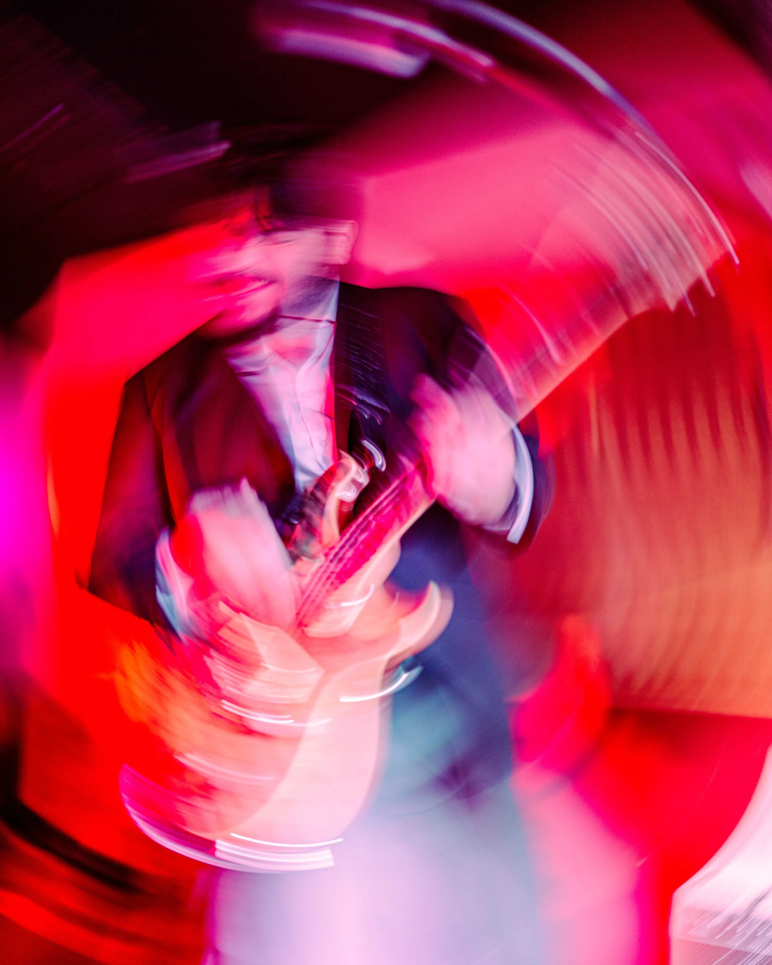 A person playing an electric guitar in a room illuminated with vibrant red and pink lights, creating a swirling motion effect.