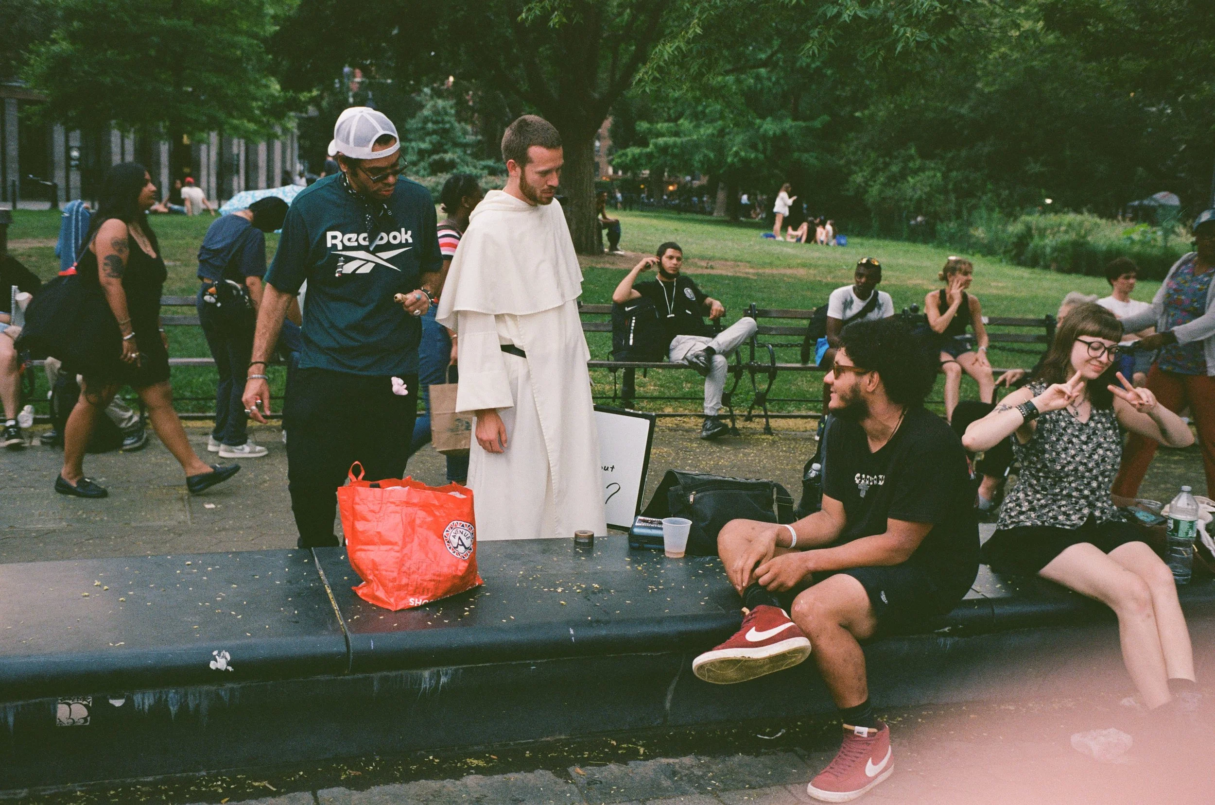 People gathered in a park, some sitting on benches and others standing, with a large tree and grass in the background.