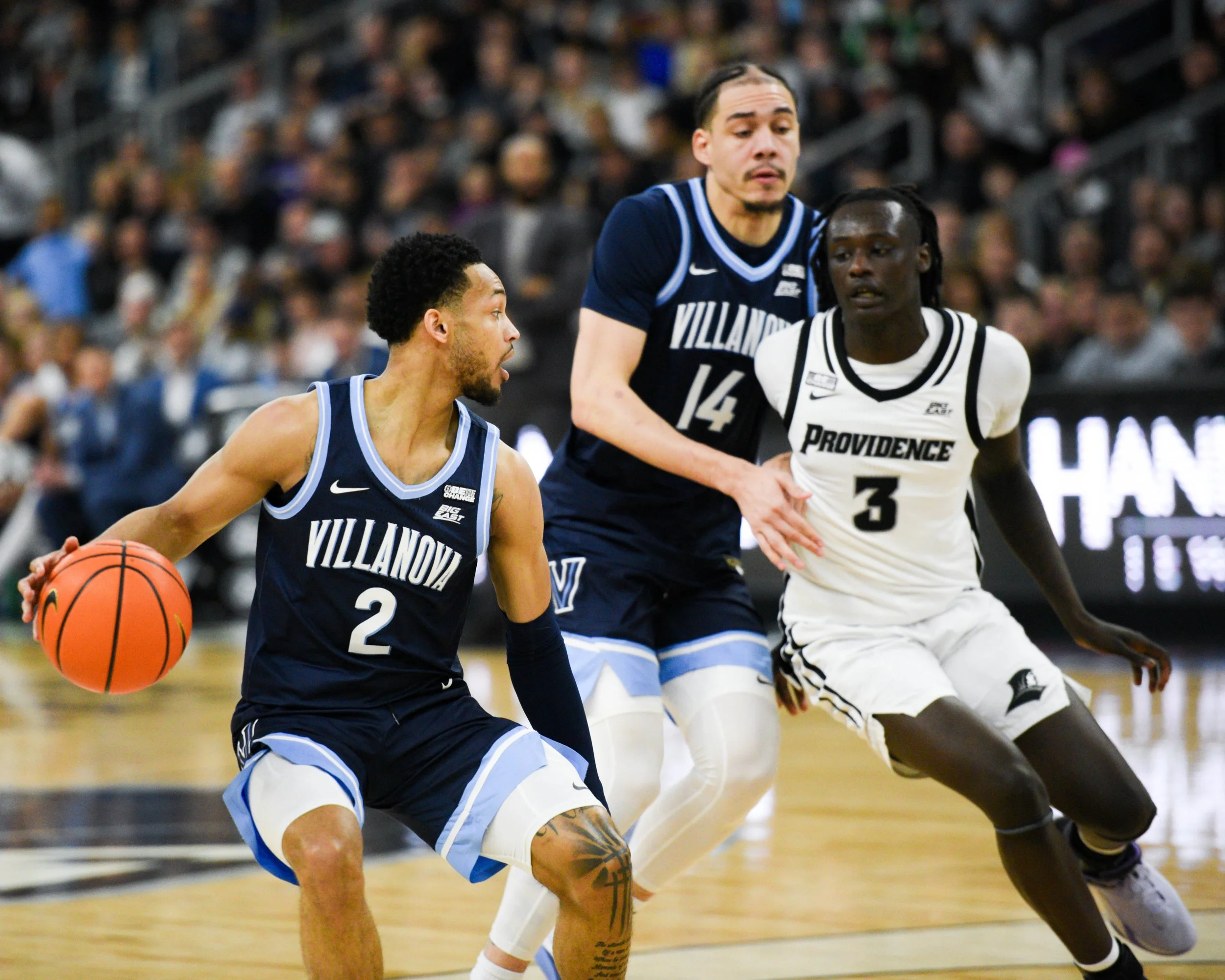 A basketball game showing three players in action. The player wearing number 2 from Villanova is dribbling the ball, while the players wearing numbers 14 and 3 from Villanova and Providence are defending.