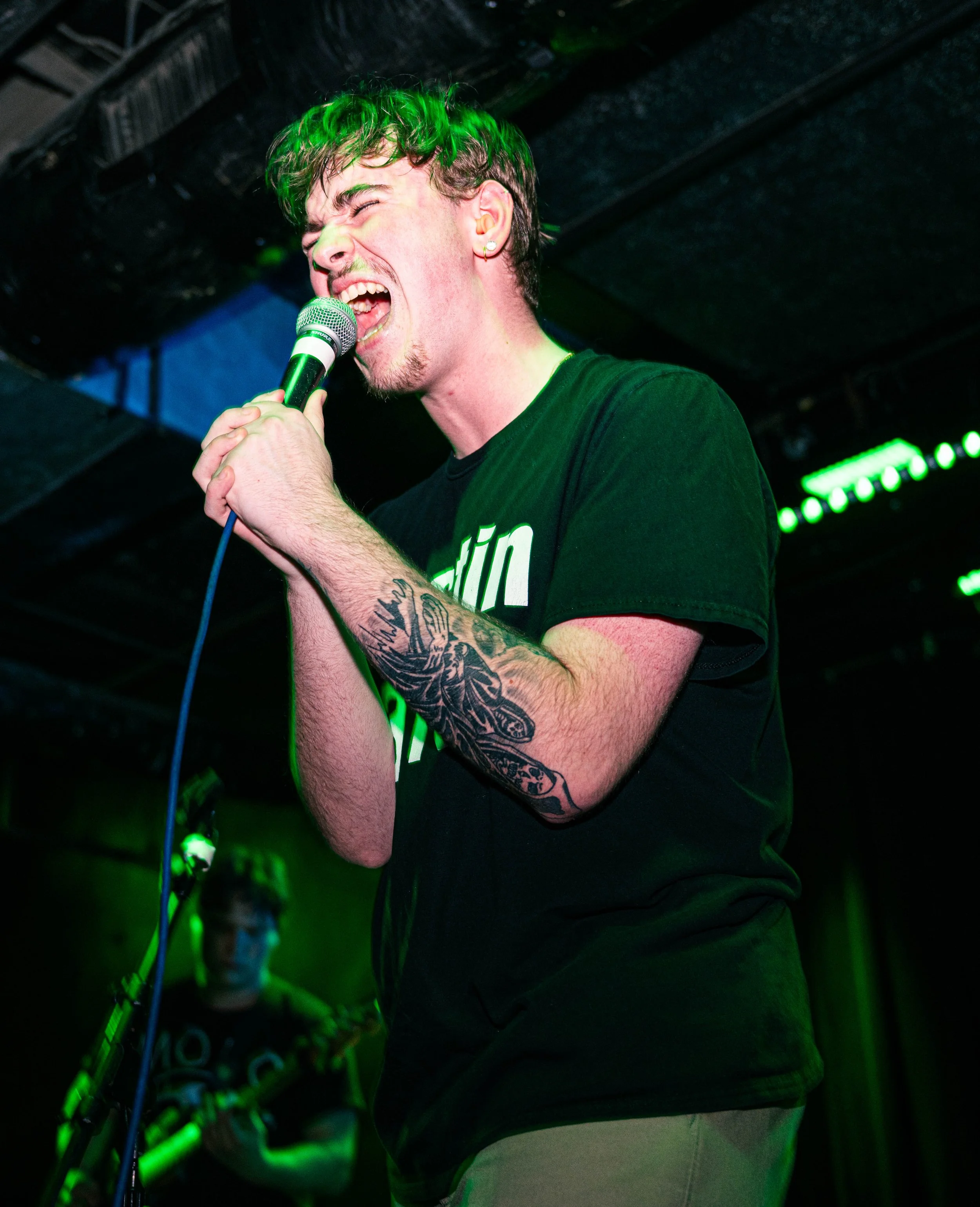 Male singer passionately performing with microphone, tattooed arm, in a music venue with green lighting, audience in background.