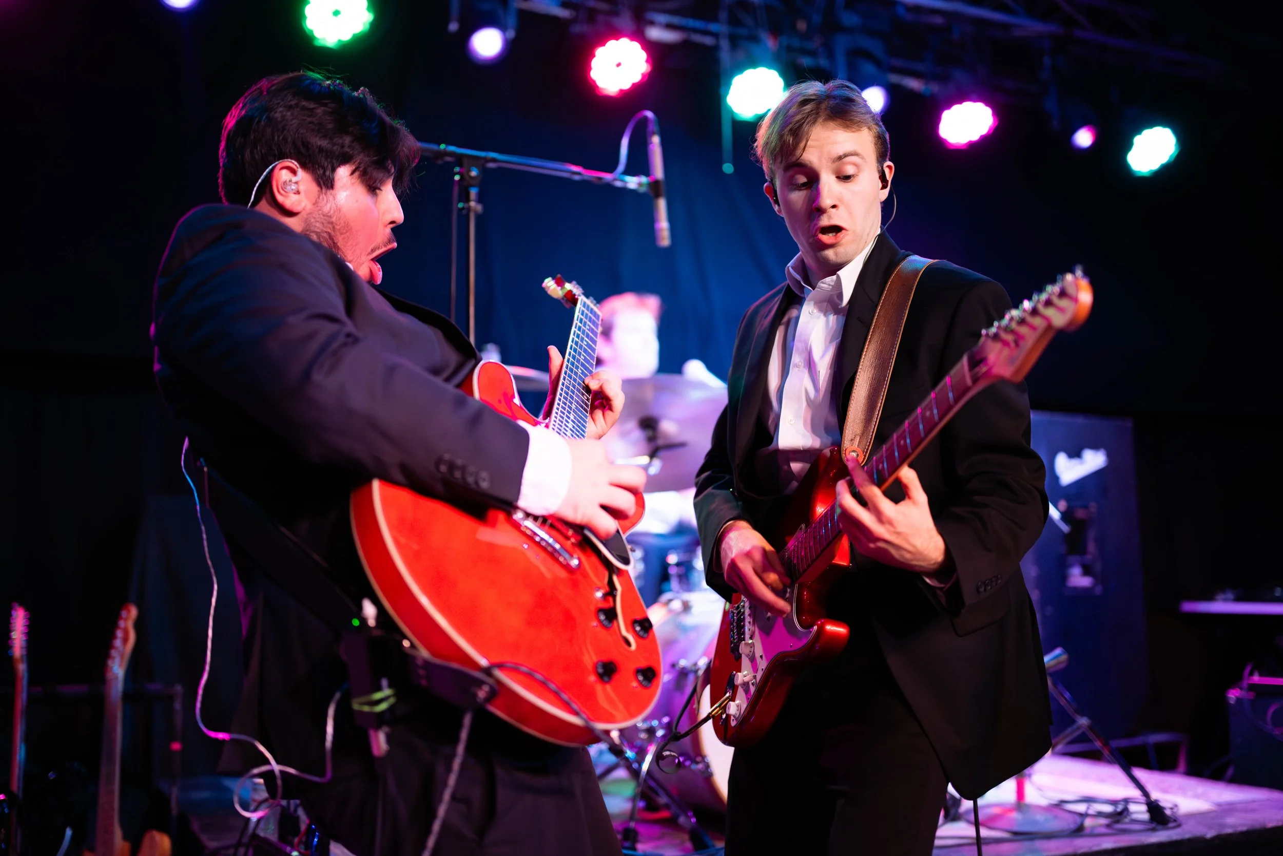 Two male musicians in suits playing electric guitars on stage during a concert, with colorful stage lights overhead.