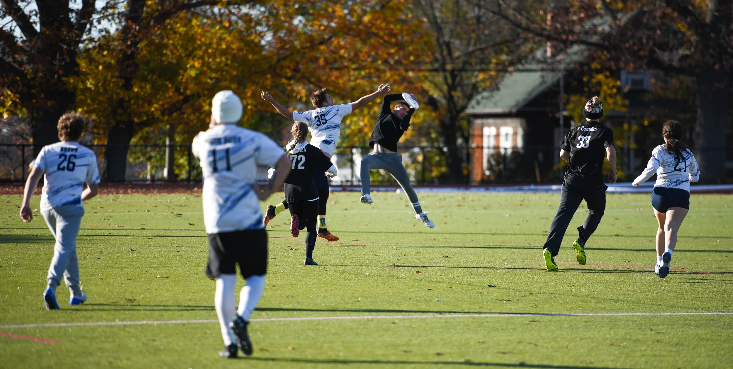 Youth soccer game on a grassy field with players in white and black jerseys, some jumping to catch the ball, surrounded by trees with fall foliage.