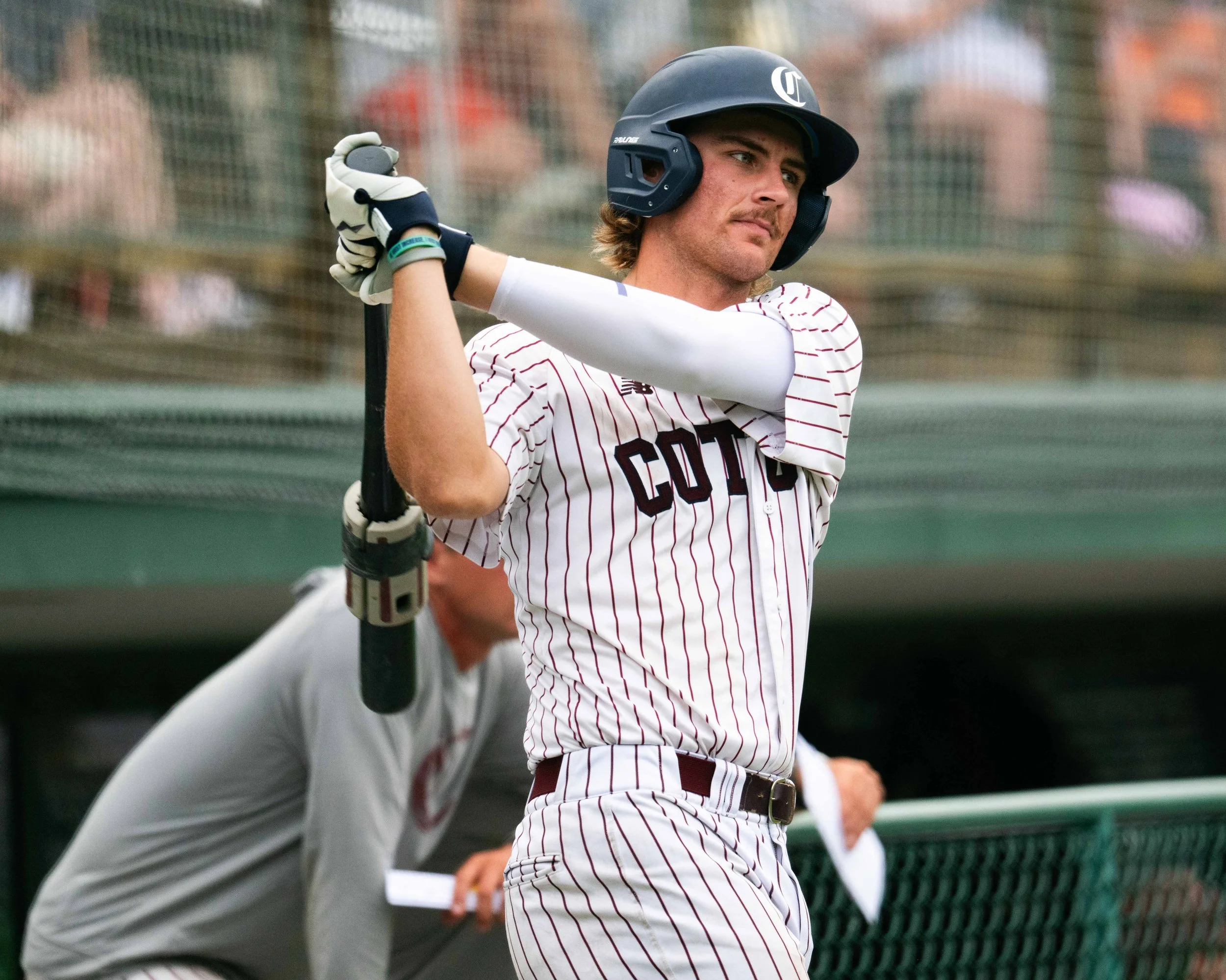A young male baseball player wearing a helmet and uniform is holding a bat, preparing to swing during a game. There is a coach or teammate behind him.