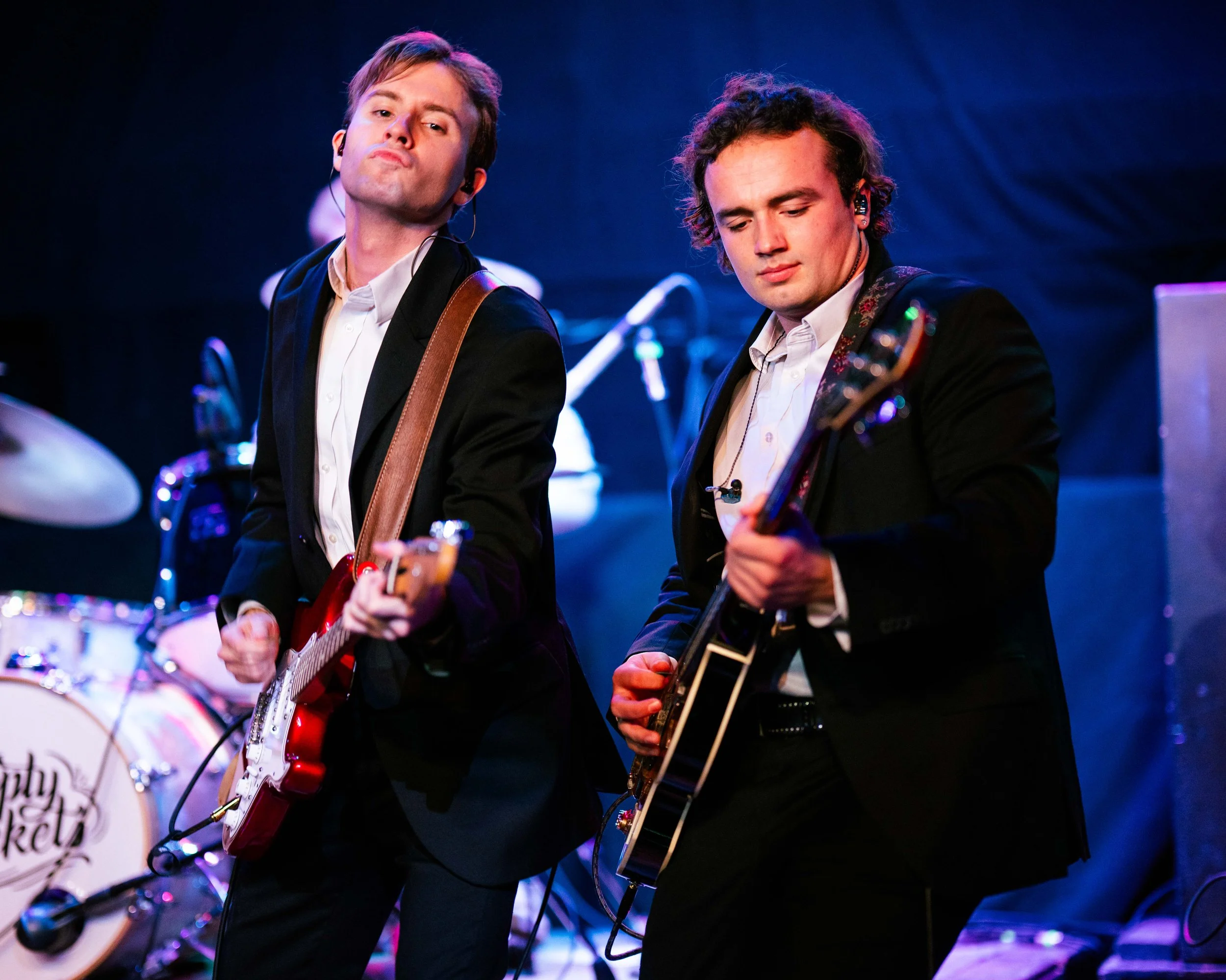 Two men playing guitars on stage during a live music performance, with drums in the background.